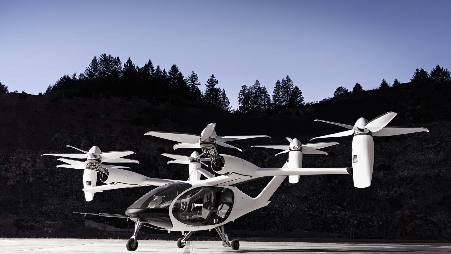 Joby electric air taxi, with six large rotors and a transparent canopy, parked on a concrete surface with forested hills and a clear sky in the background.