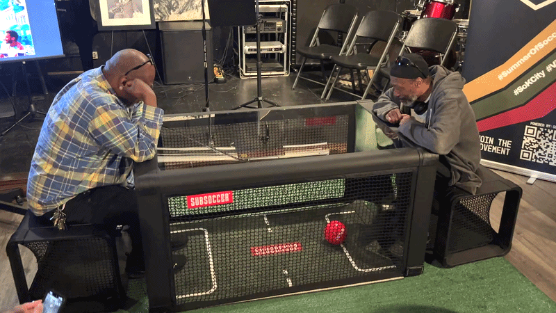 Two men sit at a transparent table kicking a red soccer ball until one person scores and smiles.