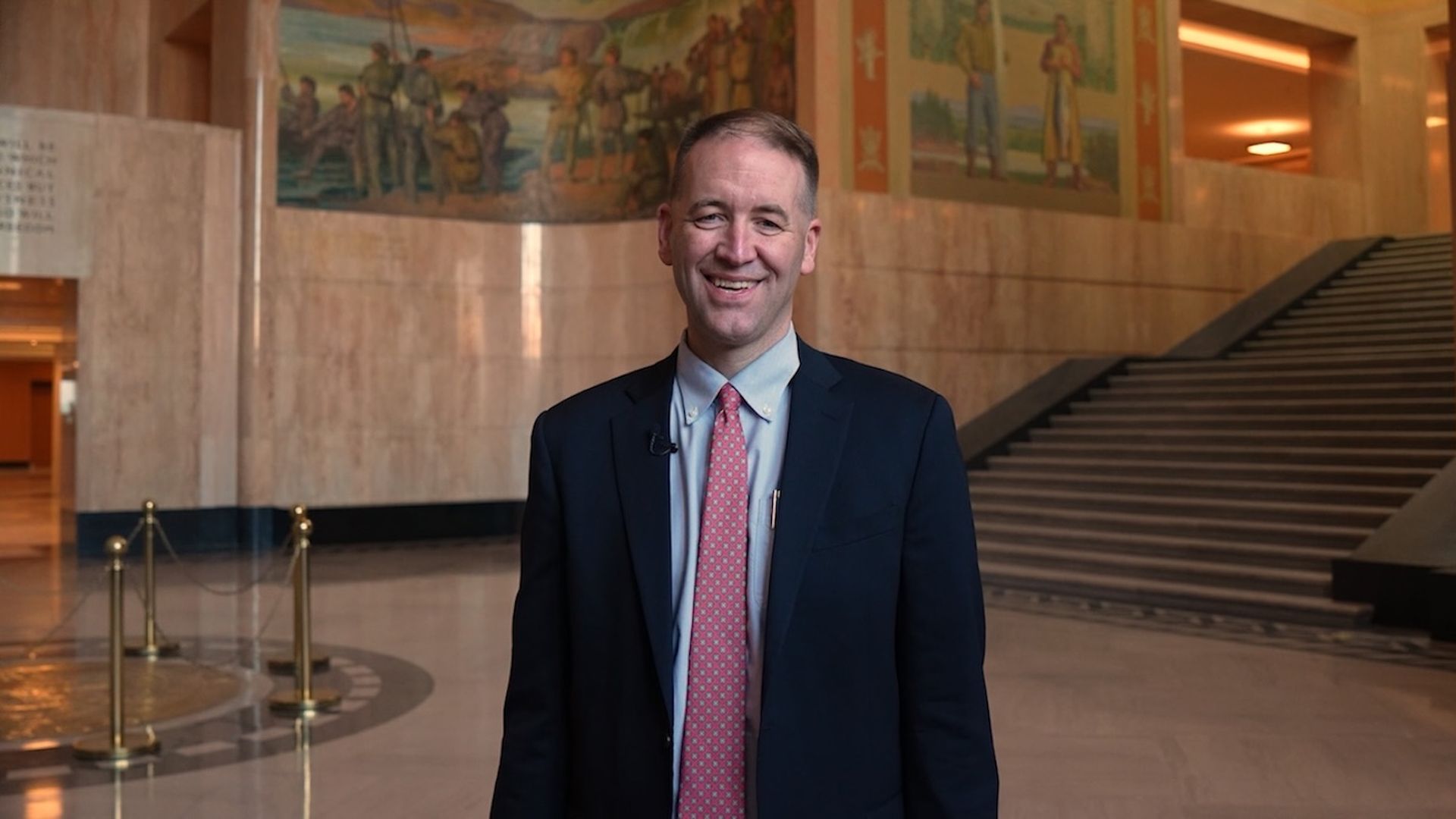 Smiling man in a navy suit, light blue shirt, and pink patterned tie stands in a marble lobby with a wide staircase and murals behind him.