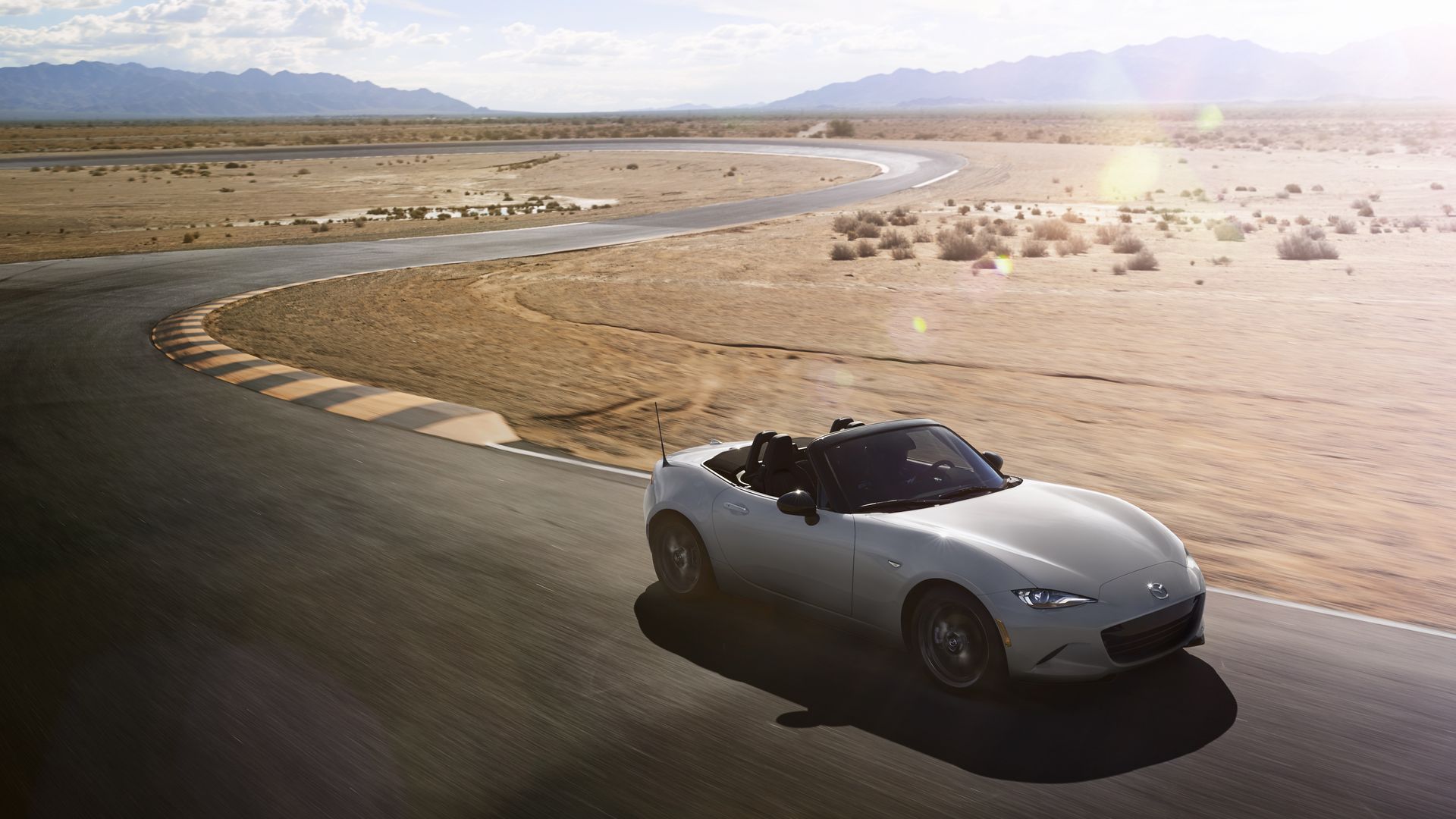 Silver Mazda MX-5 Miata convertible on a winding desert road, curving left, brown scrub and sparse shrubs, distant blue mountains, bright sun with lens glare on the right.