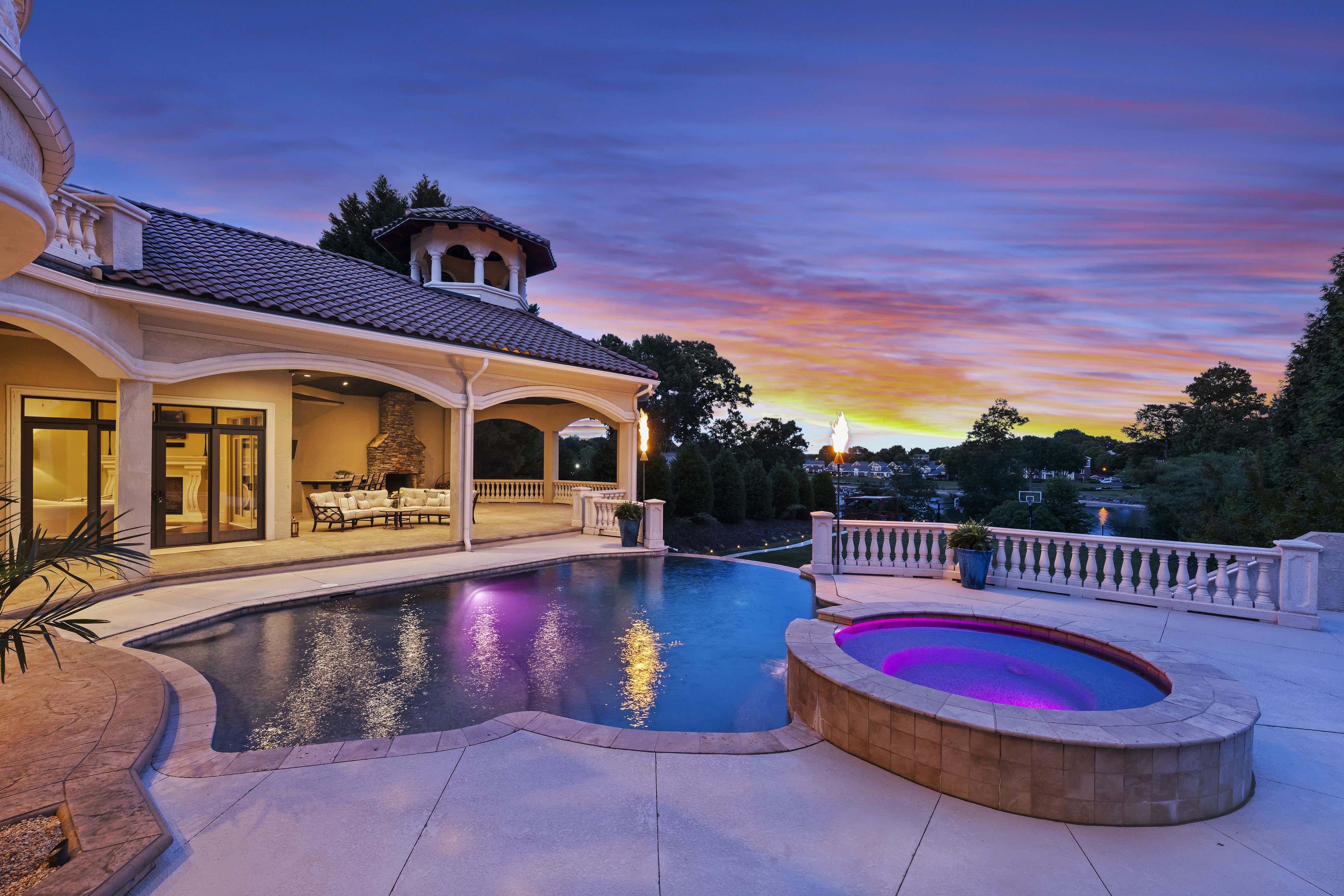 Luxurious backyard at sunset: cream house with tiled roof, arched porch and outdoor seating, beside a curved pool with purple lighting; a torch glows under a purple-blue sky.
