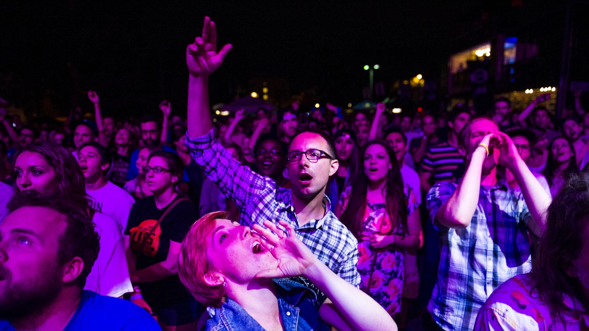 Crowd of diverse people at night, illuminated by purple and pink stage lights, some with arms raised and shouting, enjoying an outdoor concert or event.
