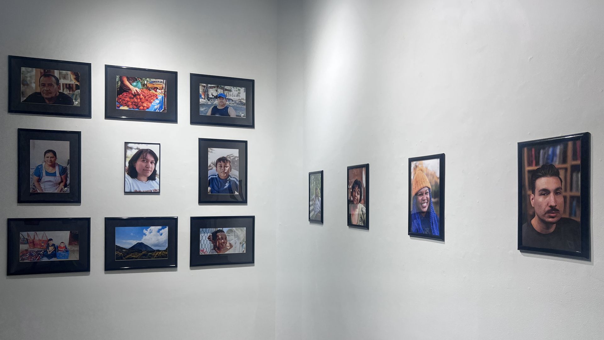 Corner of a white gallery with two walls meeting, dozens of framed portraits arranged in rows. Black frames hold diverse, candid photos of people; a large portrait dominates the right side.