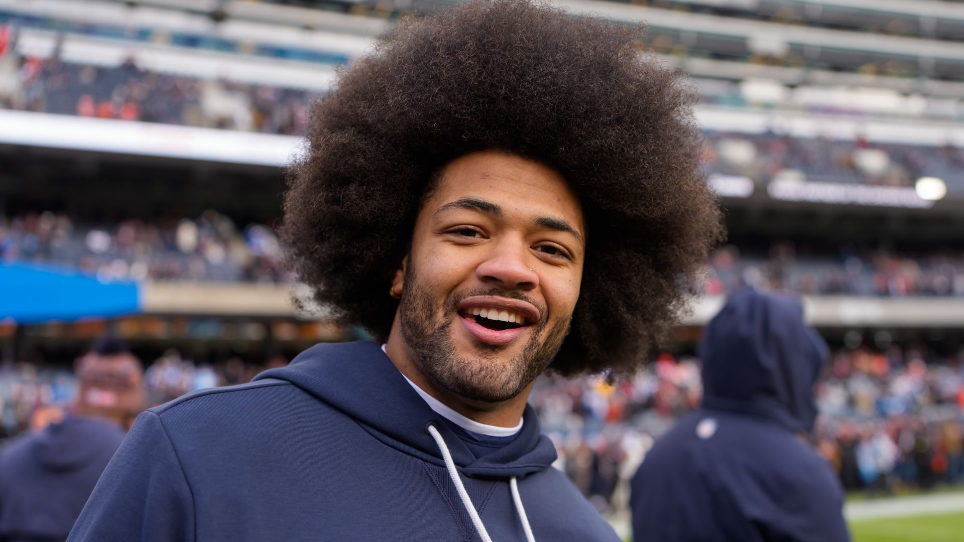Close-up of a smiling man with a large, fluffy afro wearing a navy hoodie at a stadium, with a blurred crowd and stands in the background.