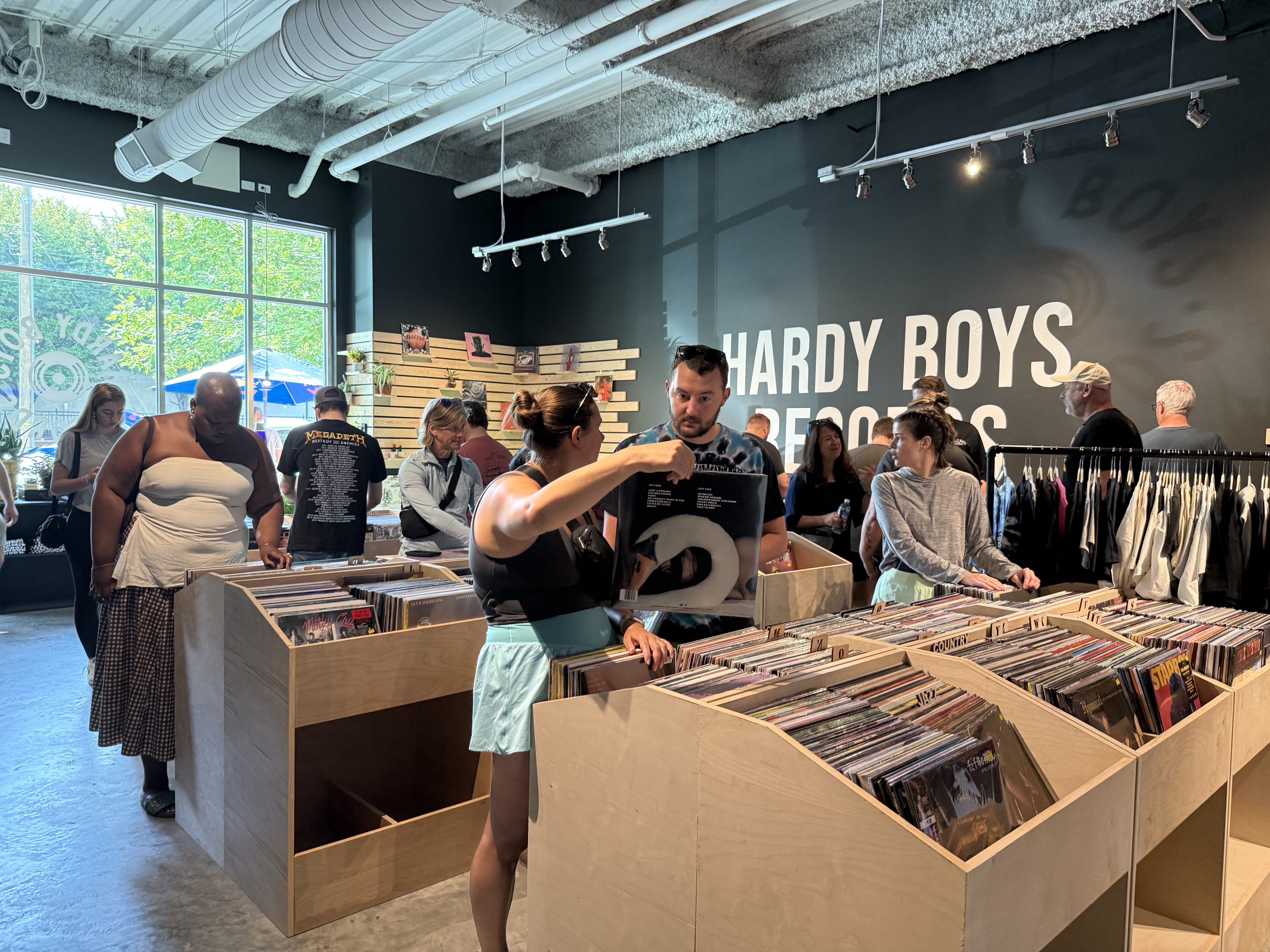 Several people browse vinyl records inside Hardy Boys Records store with black walls and natural wood bins, large windows, and a clothing rack visible on a sunny day.