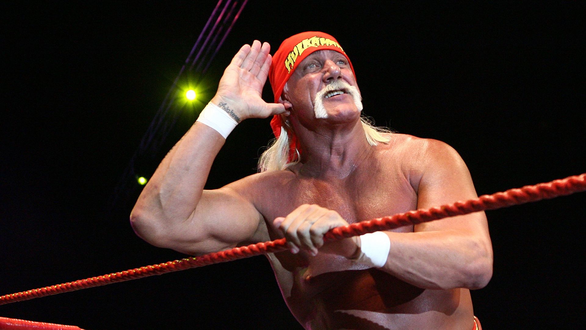 Hulk Hogan with a red bandana, white mustache, shirtless, holding red wrestling ropes, cupping hand to ear in a dimly lit wrestling arena with spotlight behind him.