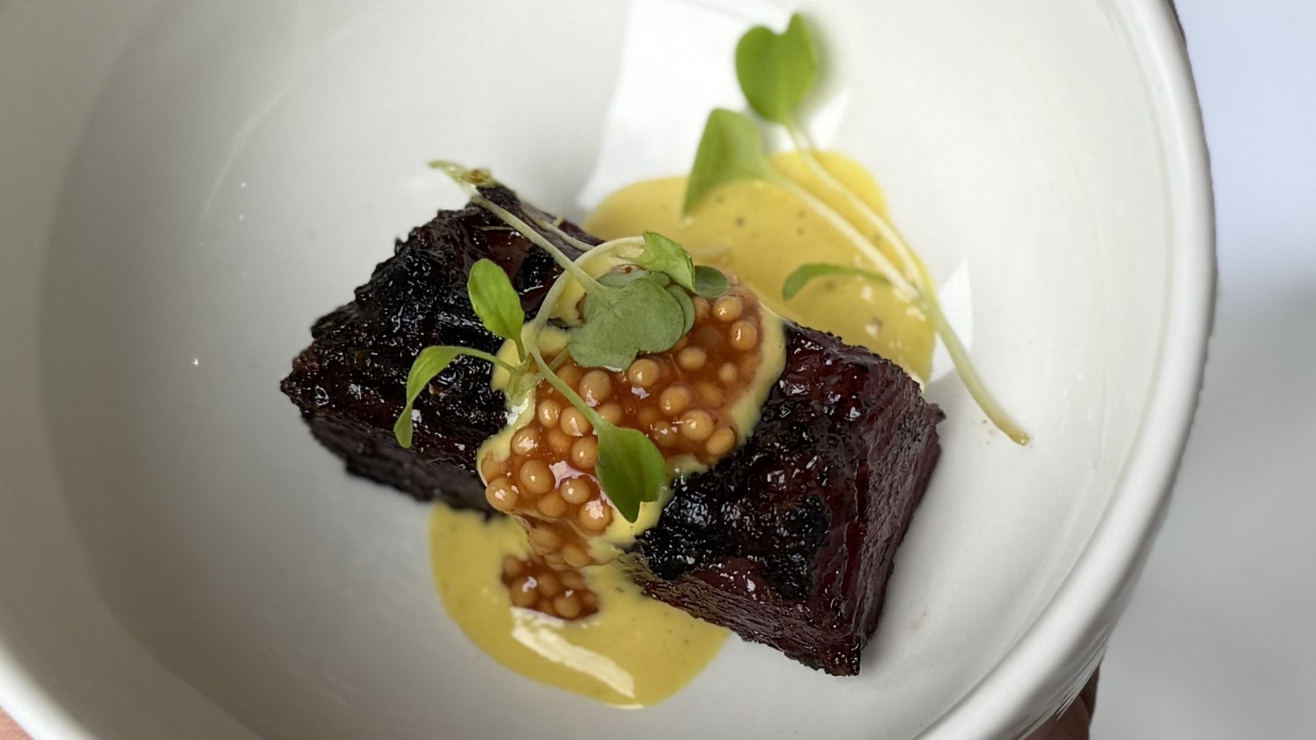 A plated dish featuring a dark, crusted meat portion topped with orange caviar-like beads, microgreens, and a yellow sauce on a white plate.