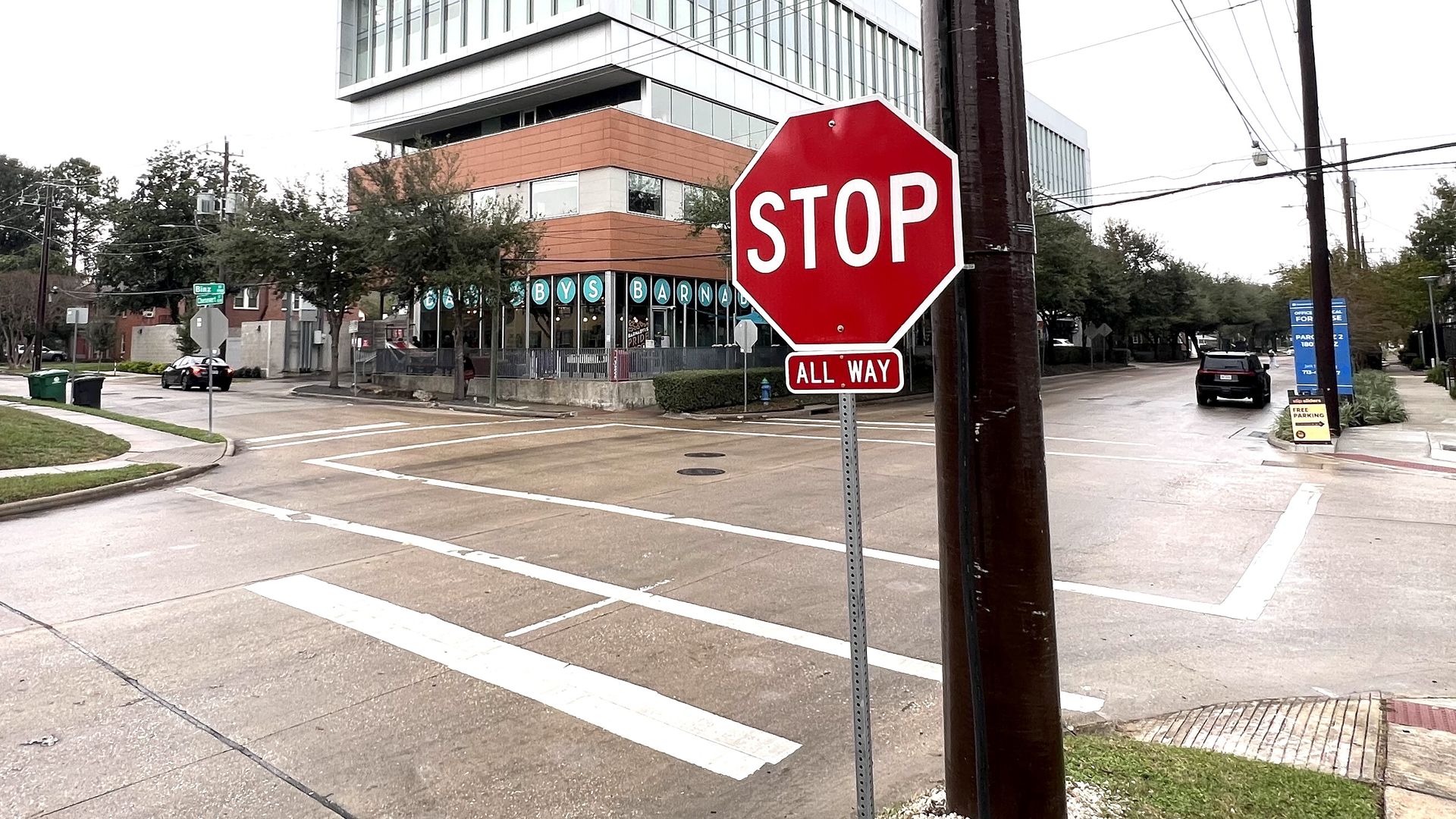 Urban street intersection on cloudy day with a red octagonal stop sign reading "STOP" and "ALL WAY," modern building with glass windows and brick facade, and utility poles with wires.