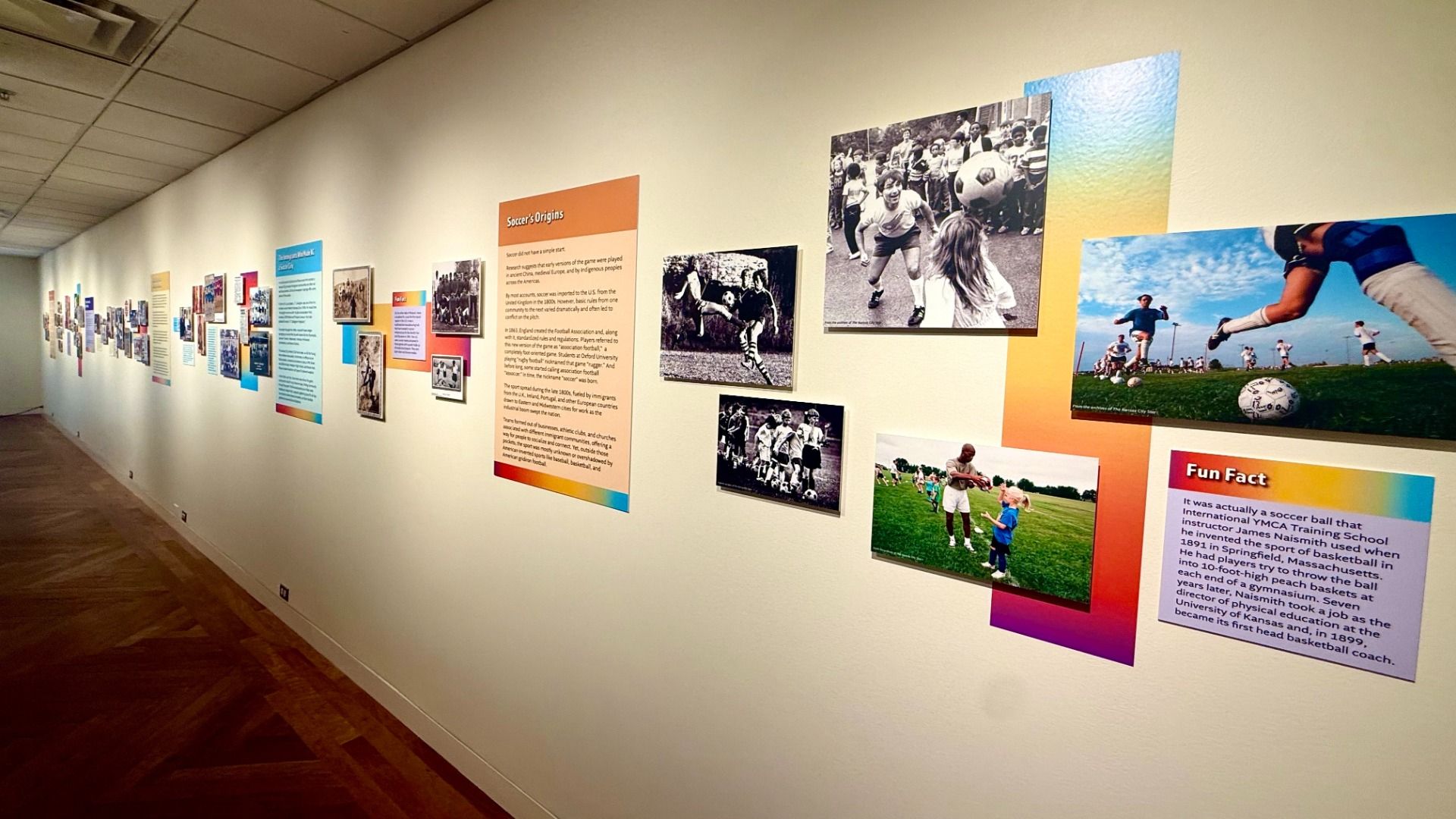 Long white gallery hallway lined with colorful exhibit panels and photos, including a large black-and-white soccer scene; smaller images and text panels adorn the wall, with a wooden herringbone floor.