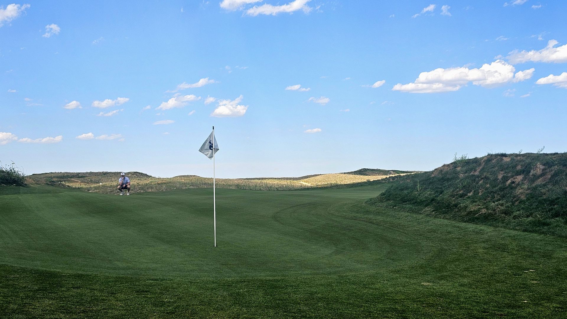 Blue sky with white clouds over a golf course green. A flagstick marks the hole in the foreground as a golfer kneels to the left, with rolling dunes in the distance.