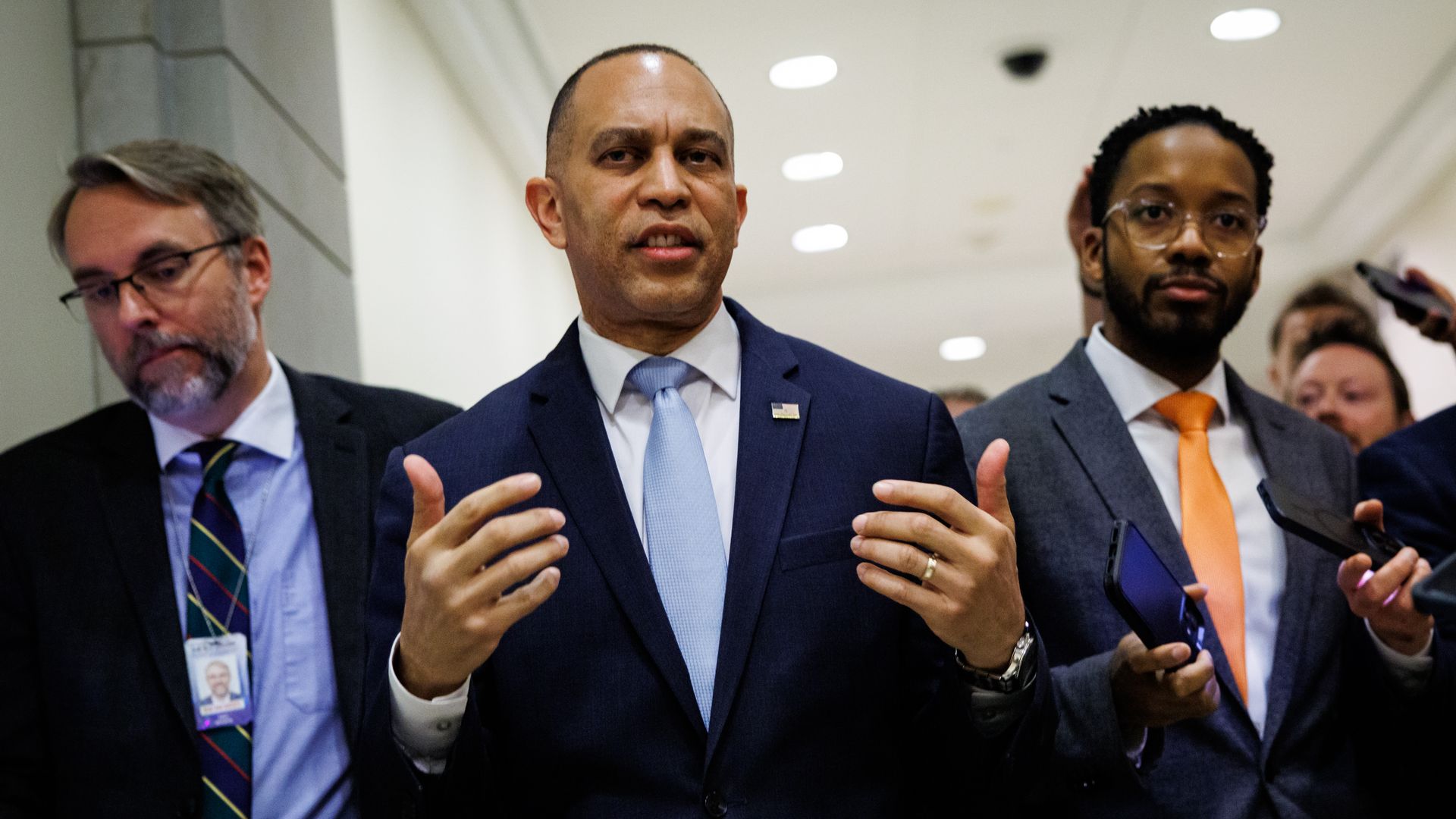 A man in a navy suit and light blue tie speaks and gestures with both hands as two other suited men stand nearby in a bright hallway.