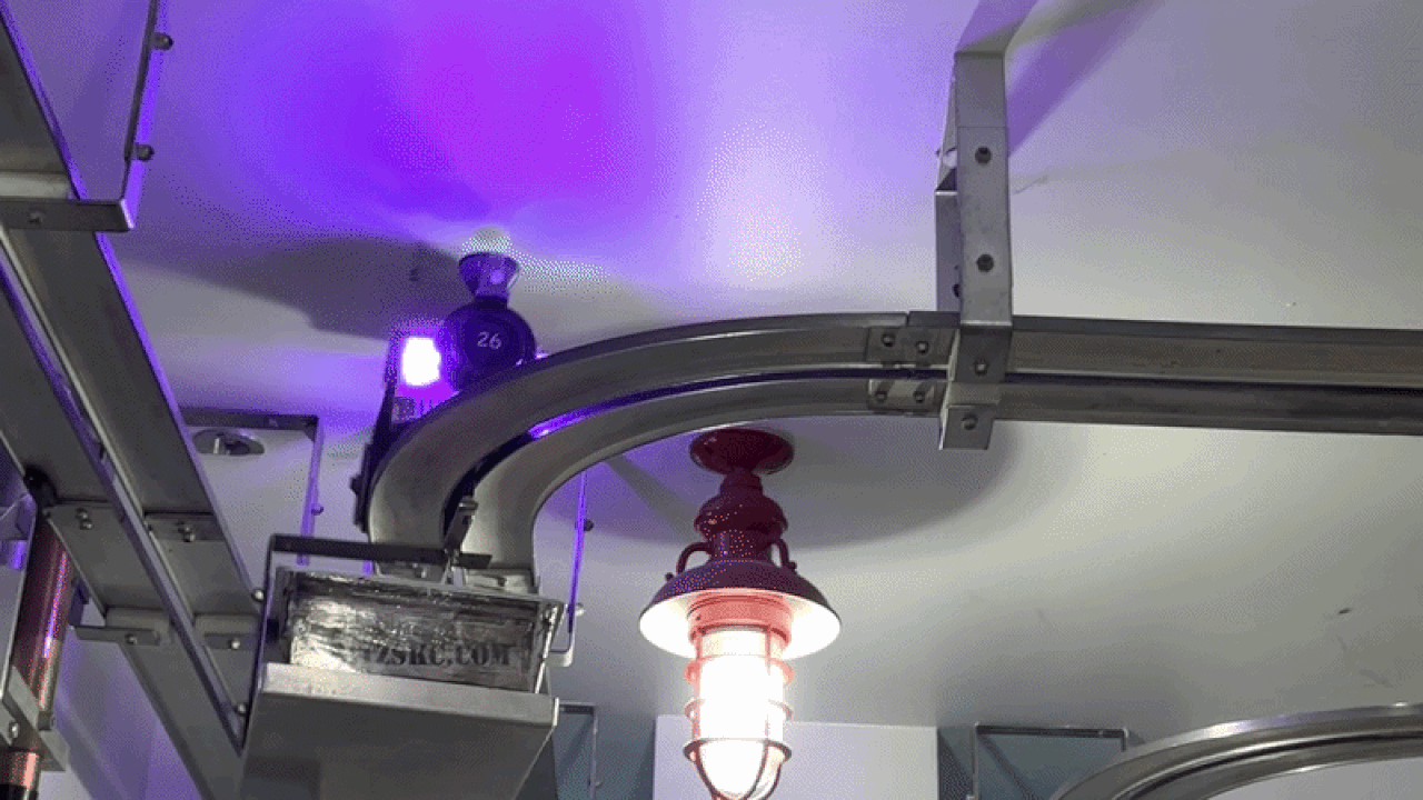 Industrial ceiling view with curved metal track, brackets, and purple lighting. A red hanging lamp centers the scene, while a small figure wearing a helmet sits on the track.