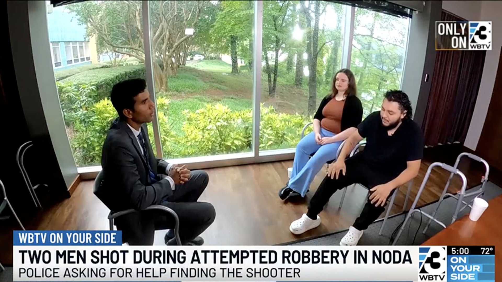 Three people sit in a room with large windows showing green trees outside. One man in a suit interviews a man and a woman, the man has a walker nearby.