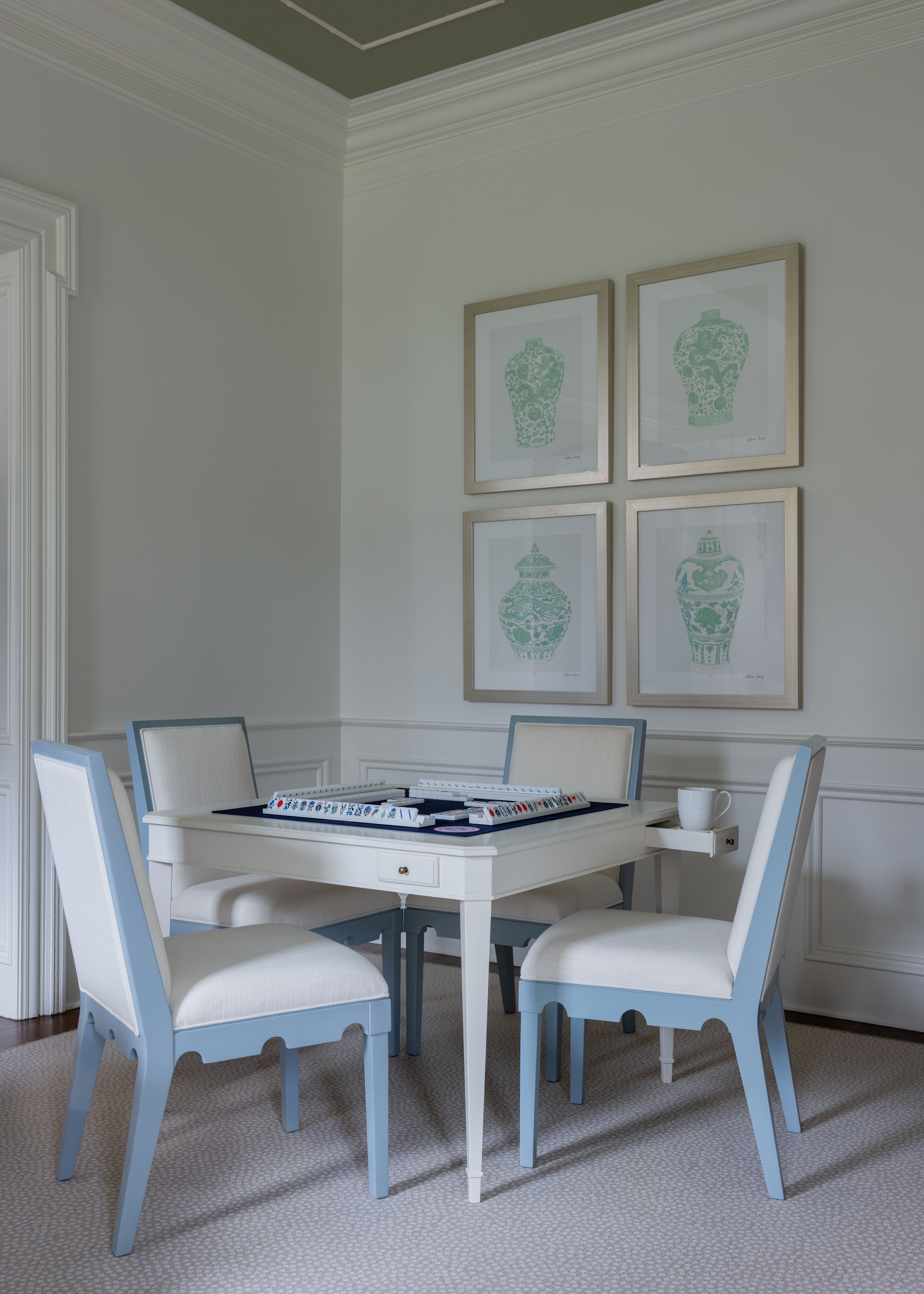 Light blue and white chairs around a white game table with mahjong tiles, set in a softly lit room with white walls and four framed aqua vase prints on the wall.
