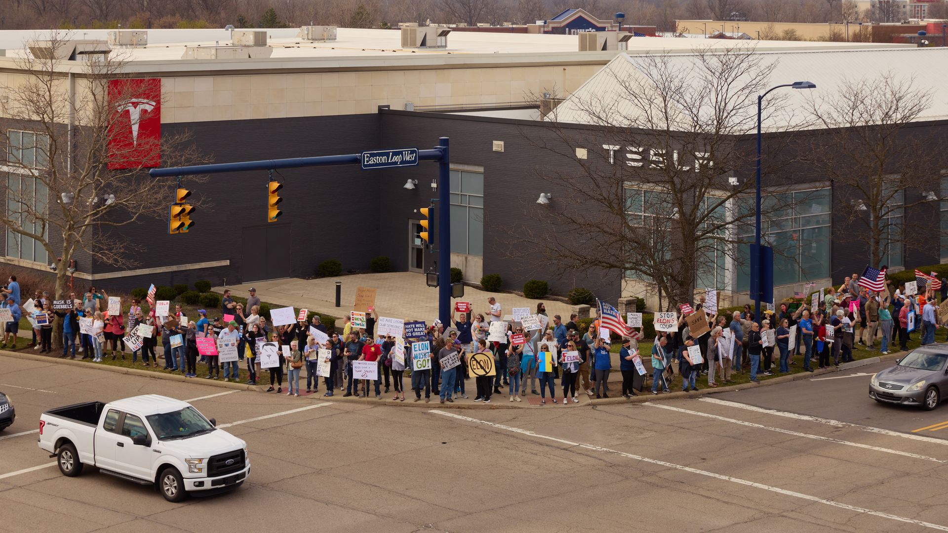 A large crowd of protestors holding signs and American flags gather outside a Tesla building on a street corner labeled Easton Loop West, with cars passing by.