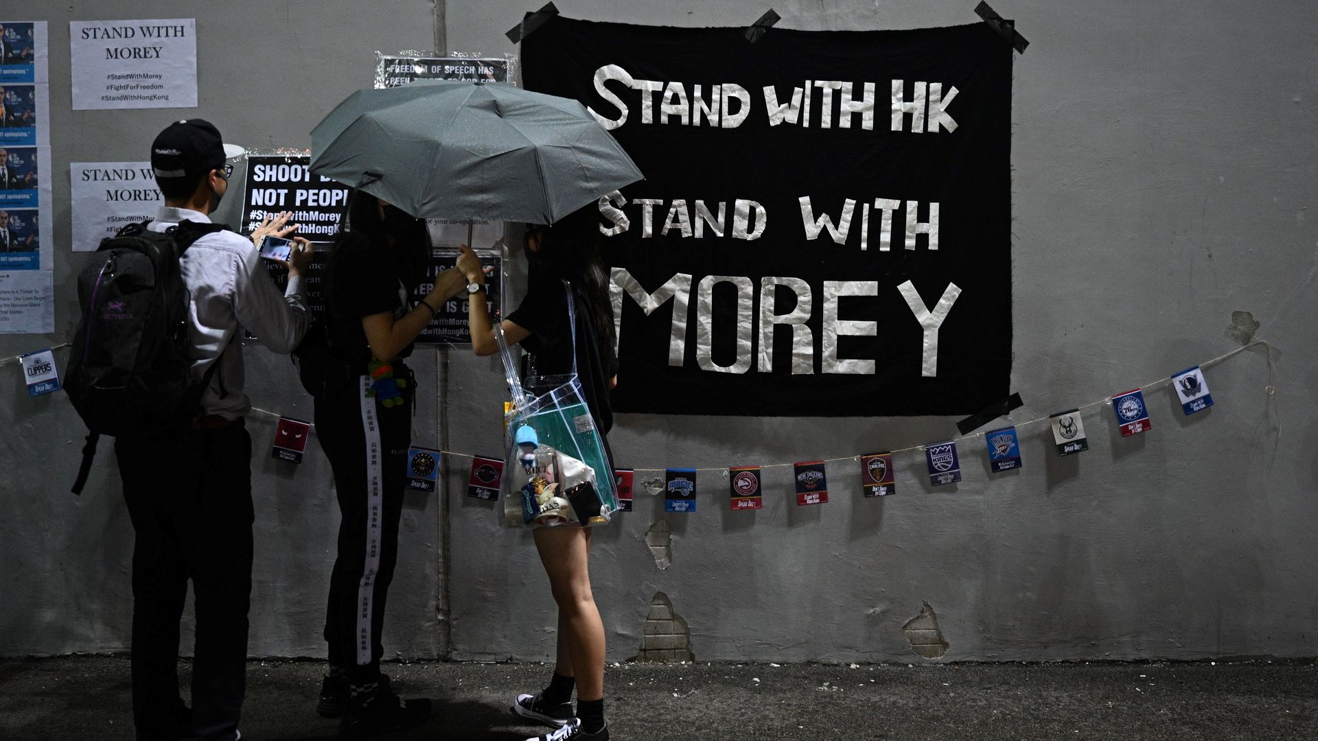 Three people in Hong Kong in front of a sign that says "stand with Morley" hanging on a wall.