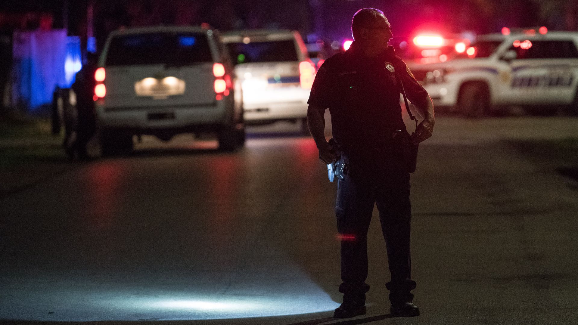In this image, a police officer stands in the middle of a darkened street at night with red police lights behind him.