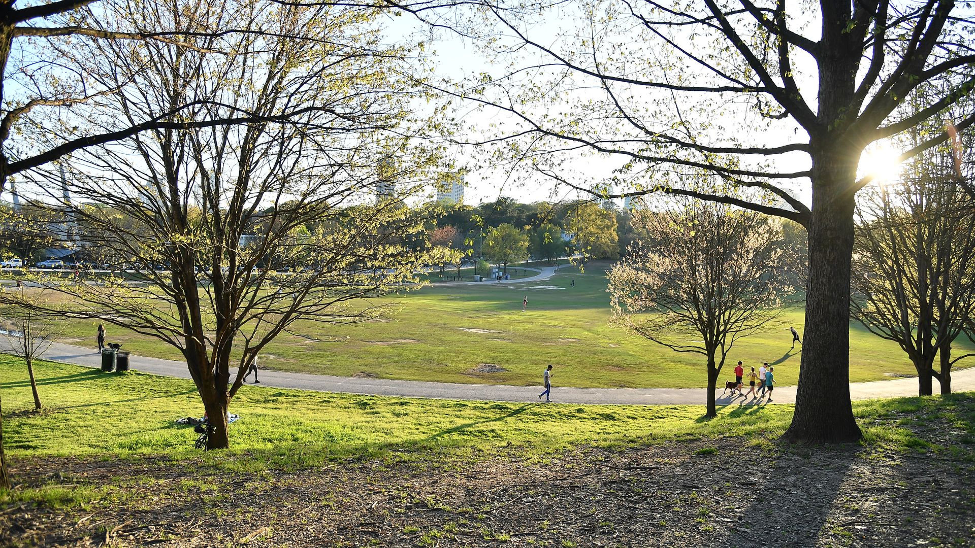 People jog in a green park with trees and a skyscraper in the background