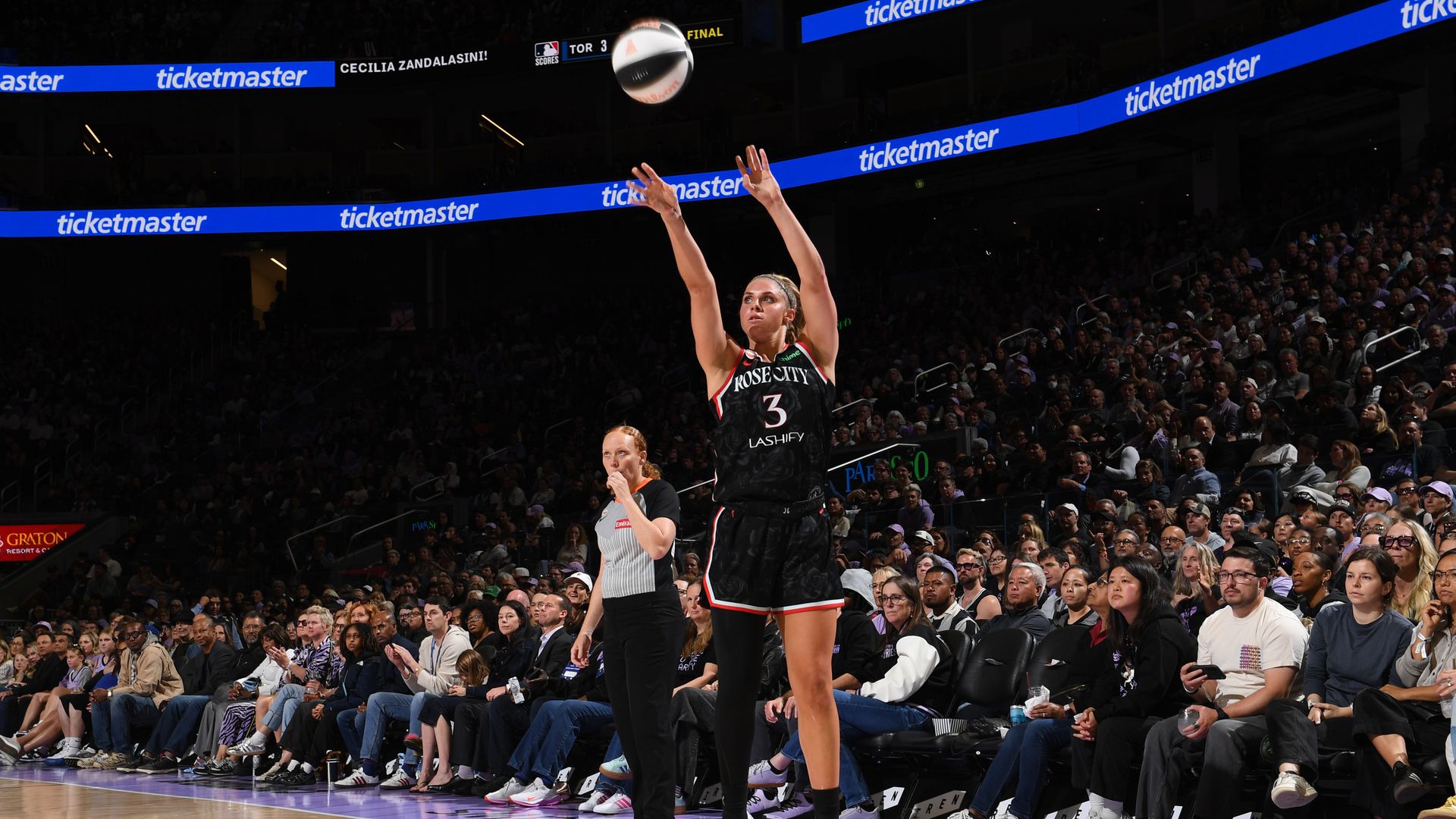 Basketball game in a crowded arena. A female player in a black Rose City jersey (number 3) jumps to block a ball as spectators watch; blue ticketmaster banners line the stands.