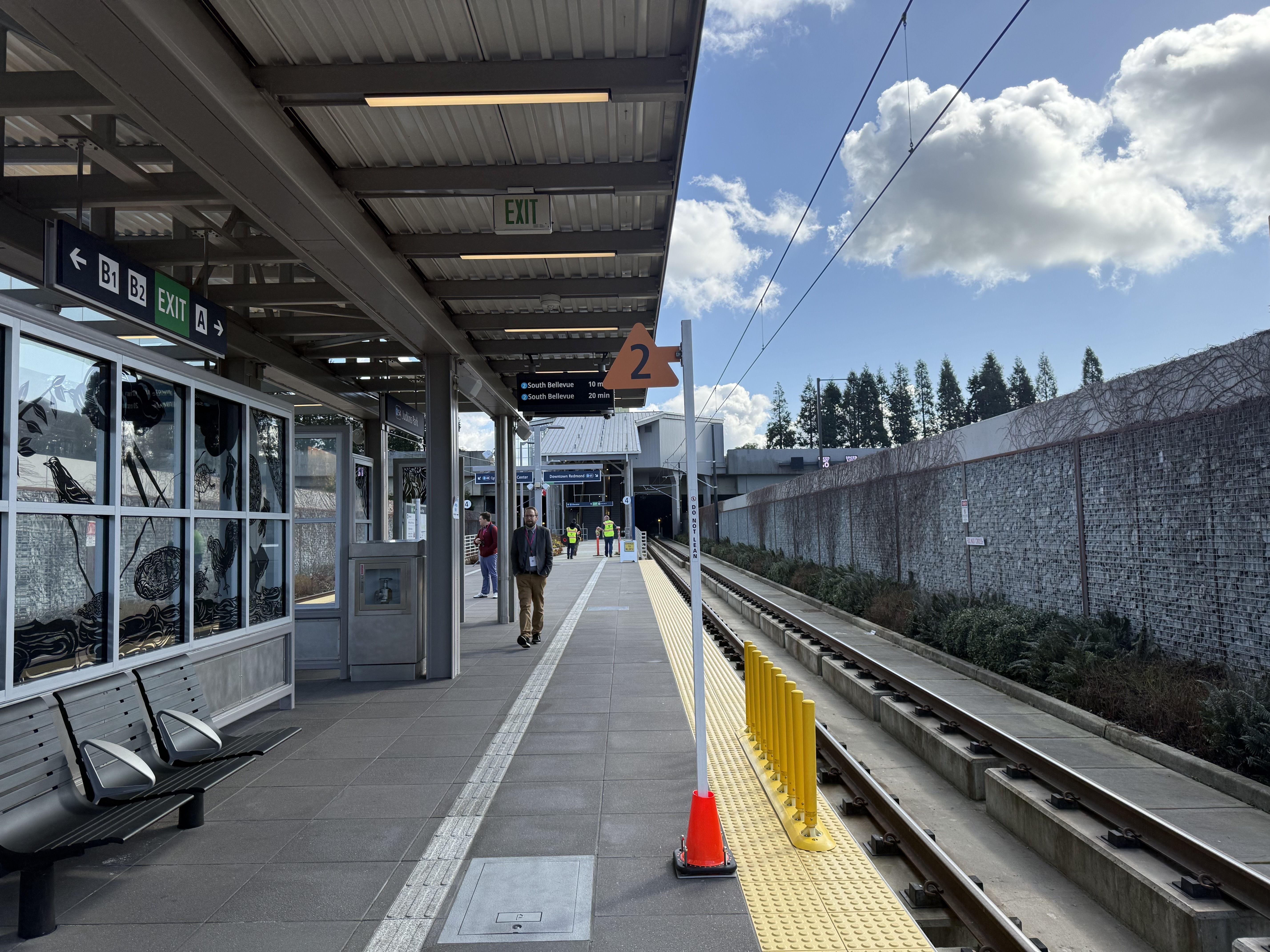 Train platform with gray benches, glass storefronts, EXIT signs, and a man walking along a yellow tactile strip beside tracks; orange cone and yellow posts under a blue sky.