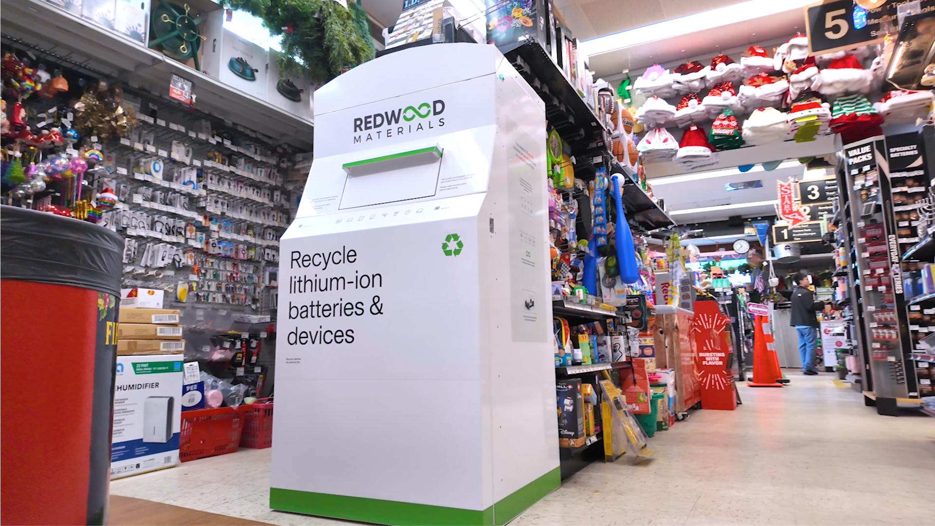 White Redwood Materials recycling bin for lithium-ion batteries and devices in a colorful store aisle with shelves of toys, costumes, and household items.