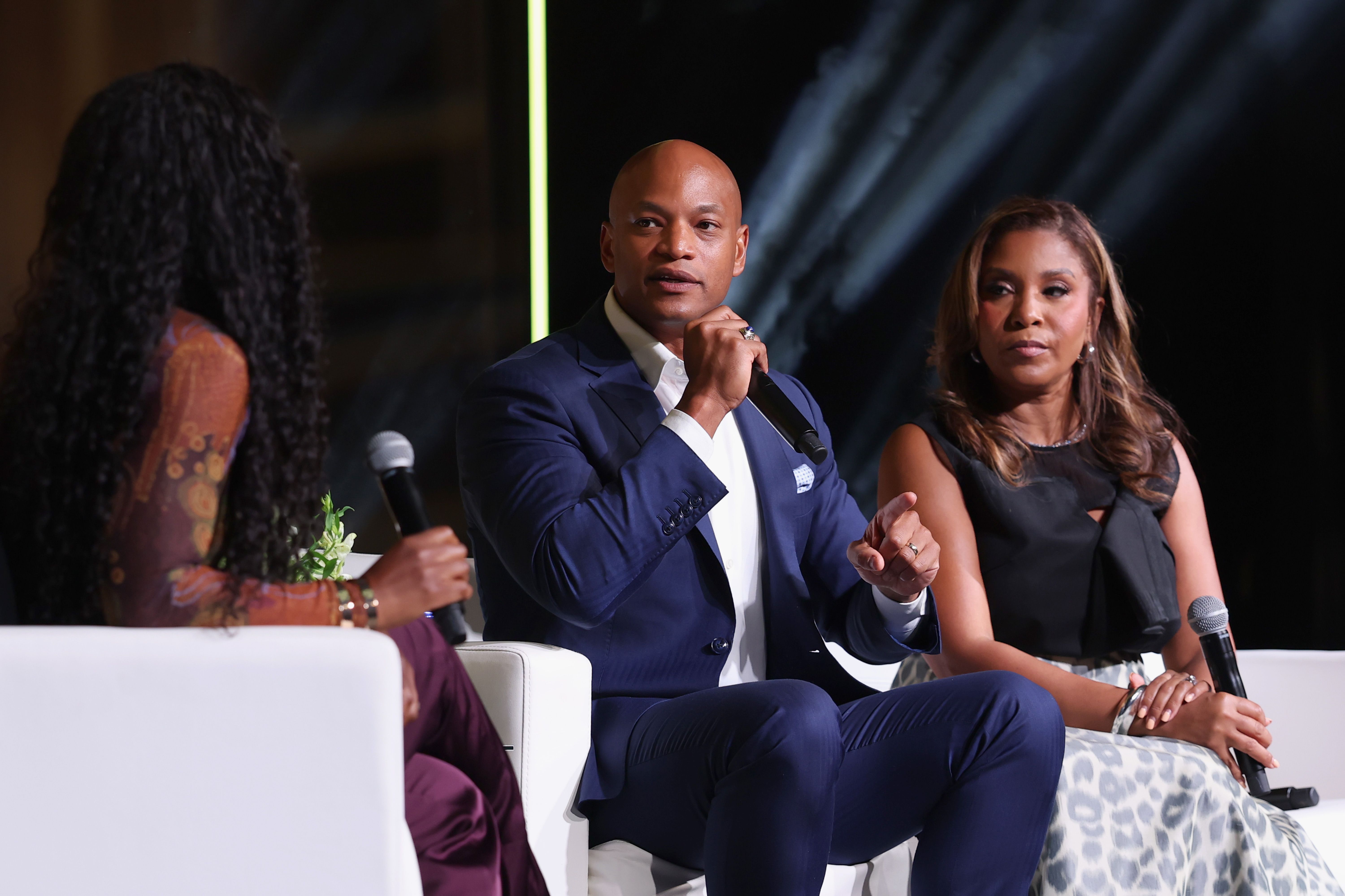 Three people in a discussion panel, two holding microphones; man in blue suit speaking, woman in black top and patterned skirt listening, third woman with curly hair partially visible.