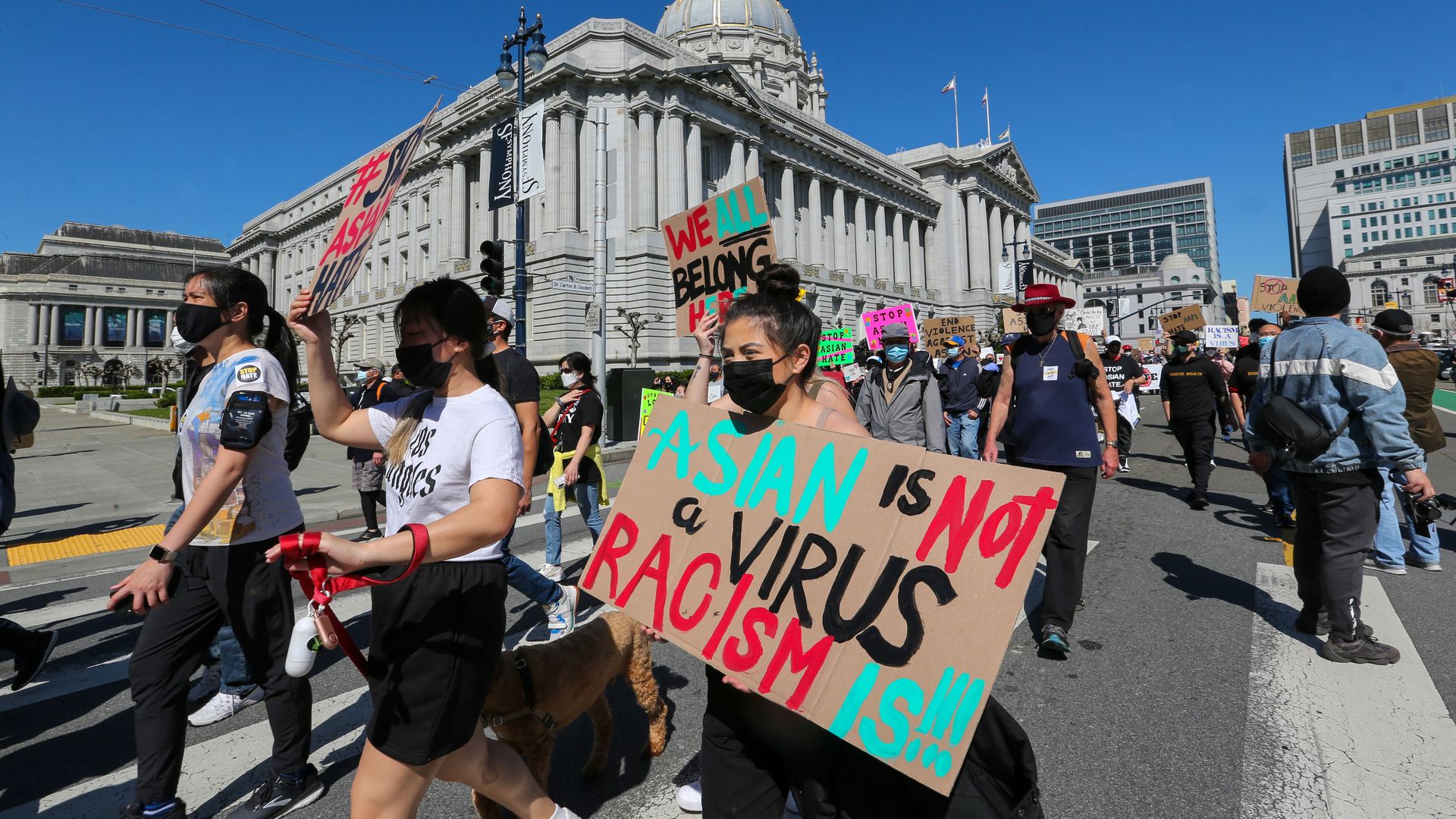 Photo of protesters marching in front of SF City Hall, with one holding a poster that says "Asian is not a virus, racism is"