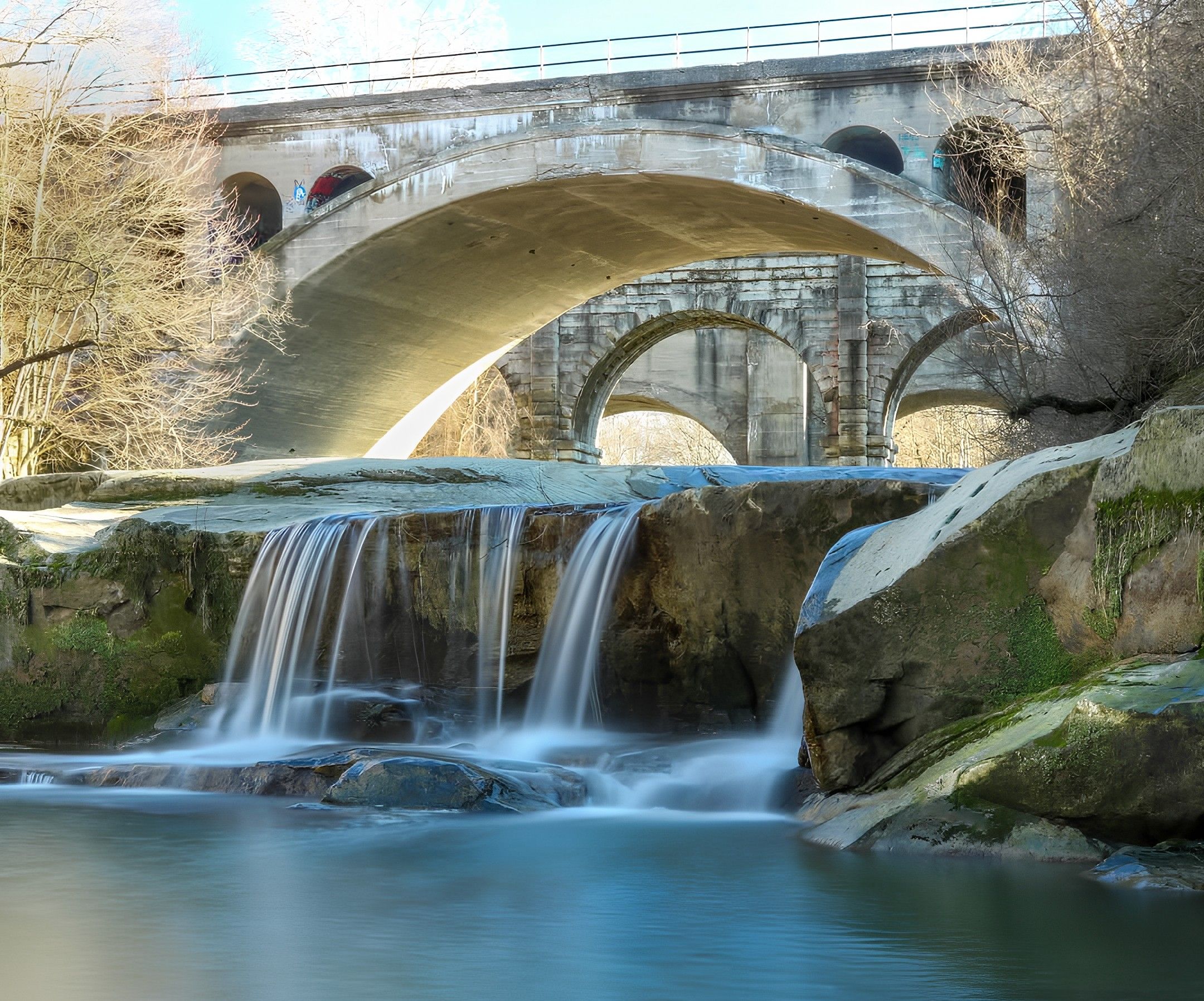 Stone arch bridge above a cascading waterfall into a blue pool; mossy rocks and leafless trees line the riverbank, with a second bridge railing in the background.