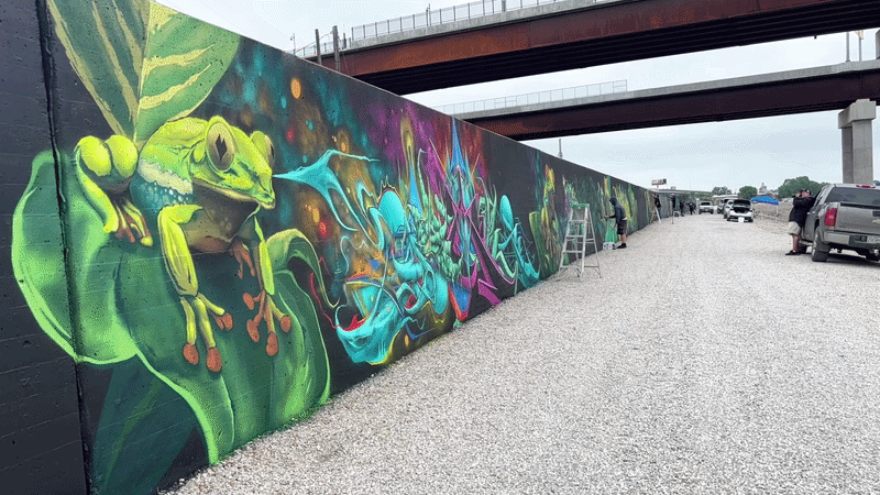 A long graffiti mural on a black wall beside a gravel road under an overpass. On the left, a bright green frog; to the right, vivid blue, purple, and pink abstract lettering. Ladders and workers.