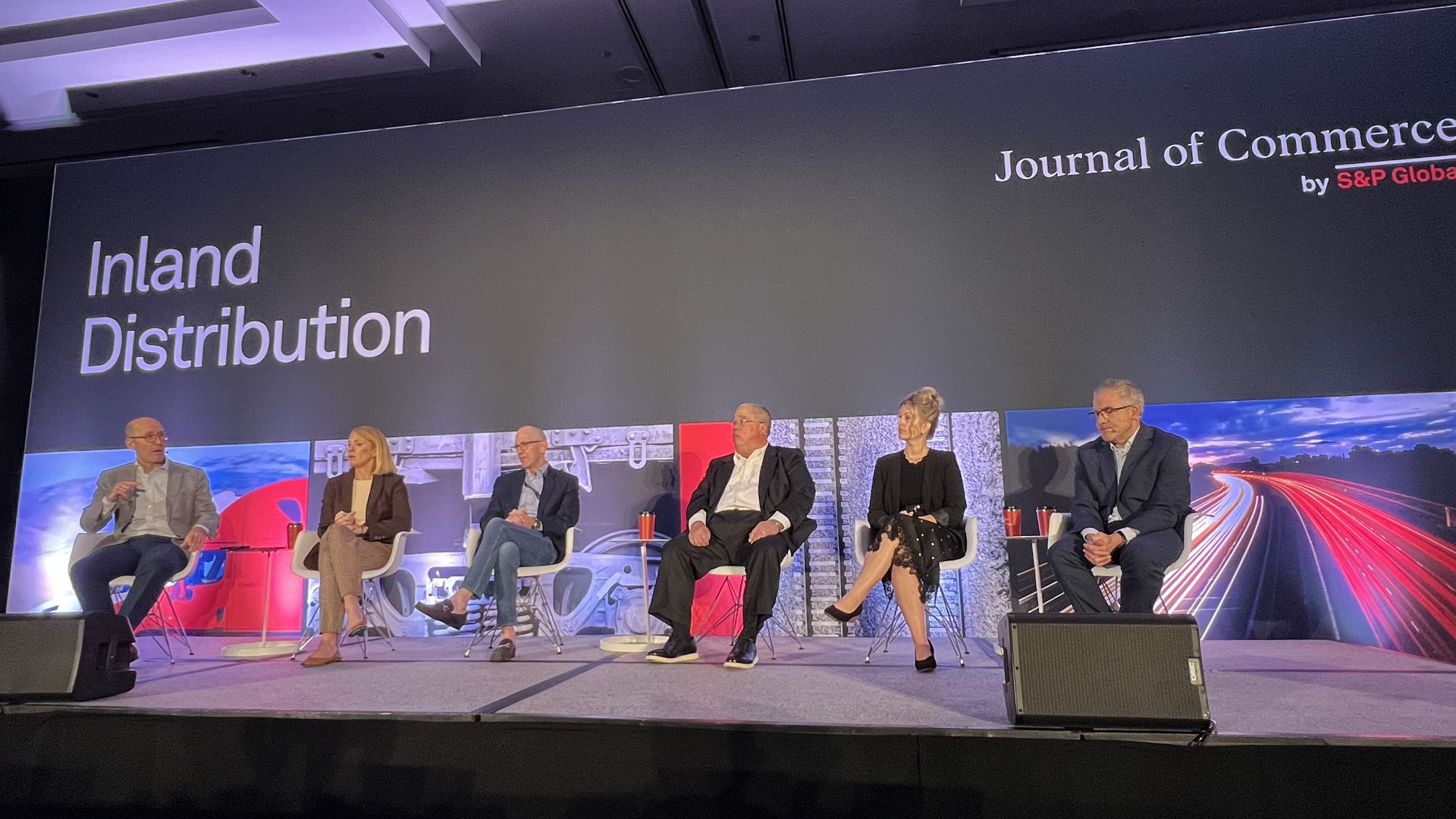 Six panelists sit on a stage in white chairs, against a dark backdrop reading "Inland Distribution" and "Journal of Commerce" by S&P Global; purple overhead lighting and colorful screens glow behind them.