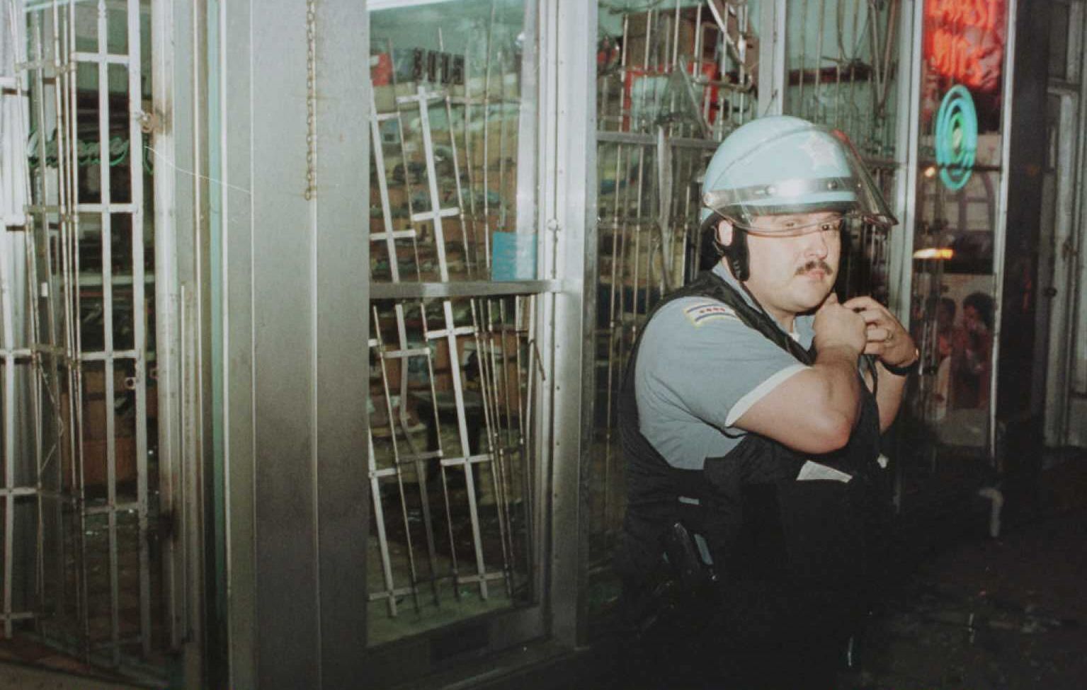 A helmeted security guard in a light blue shirt and black vest, with a clear visor, adjusts his gear beside a glass storefront with metal security grates on a dim street.