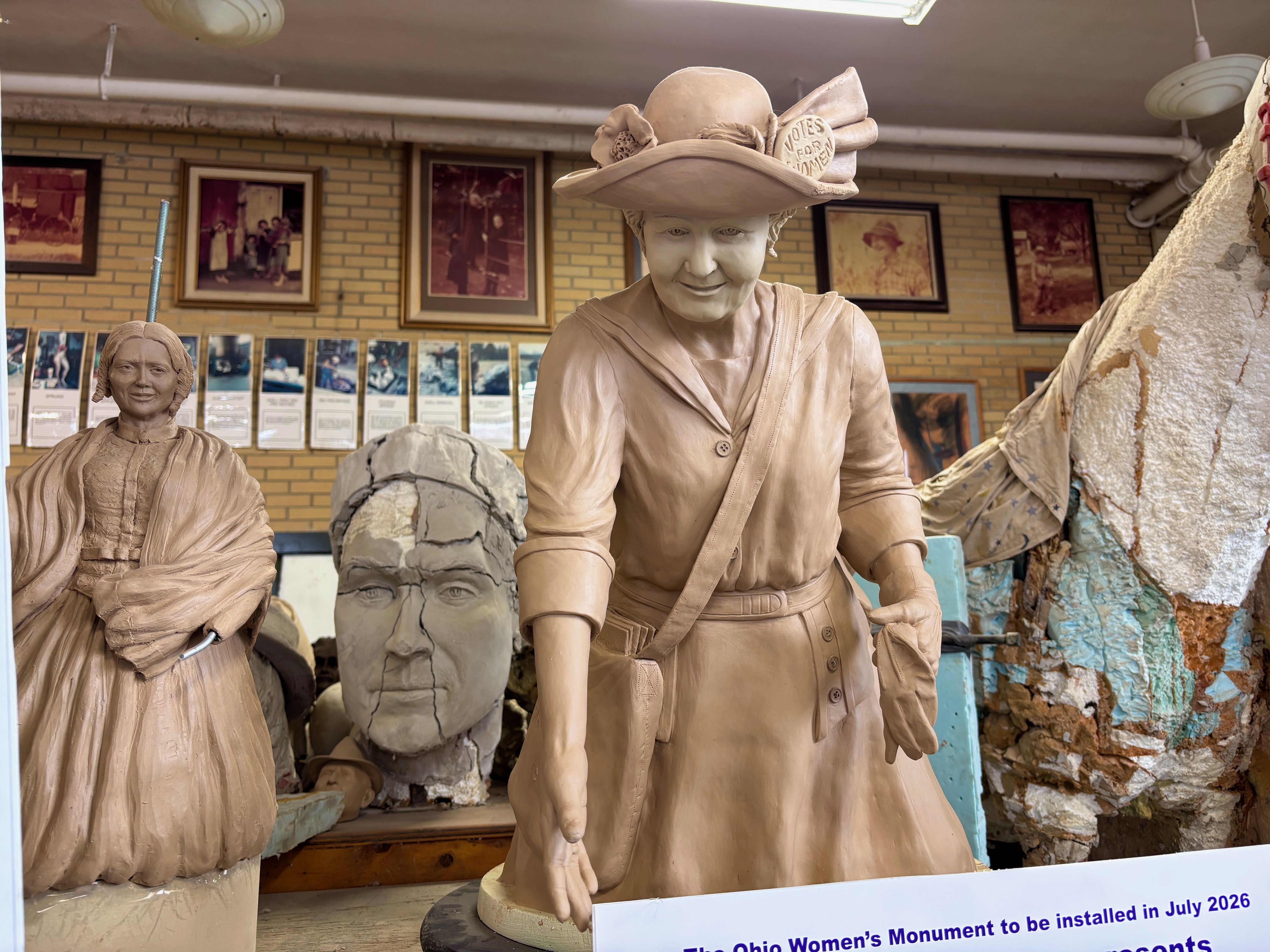 Life-size clay sculptures in a workshop: central figure with a wide-brim hat and vintage dress, a second figure to the left, cracked head behind, walls lined with framed photos.