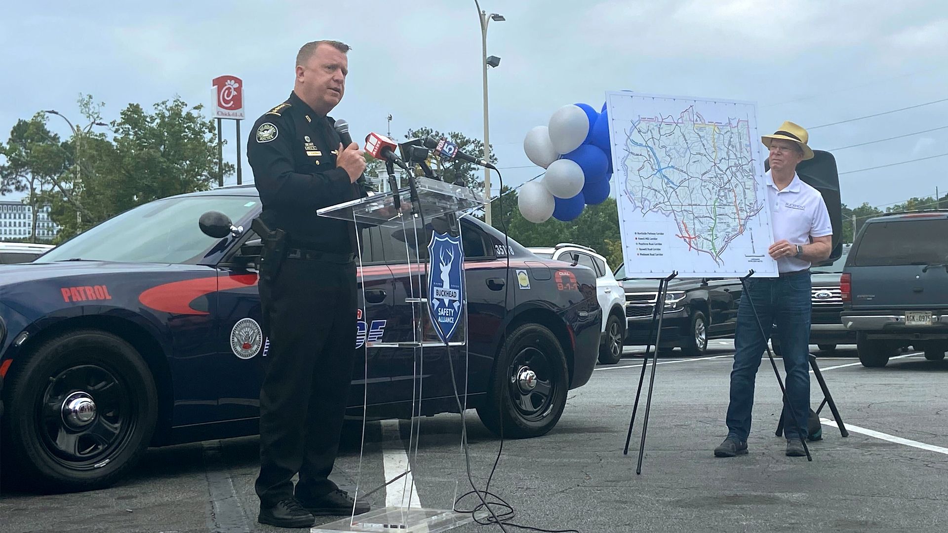 A police officer stands at a lectern in front of a cruiser while a man in a hat stands nearby holding up a map of the Buckhead police zone.