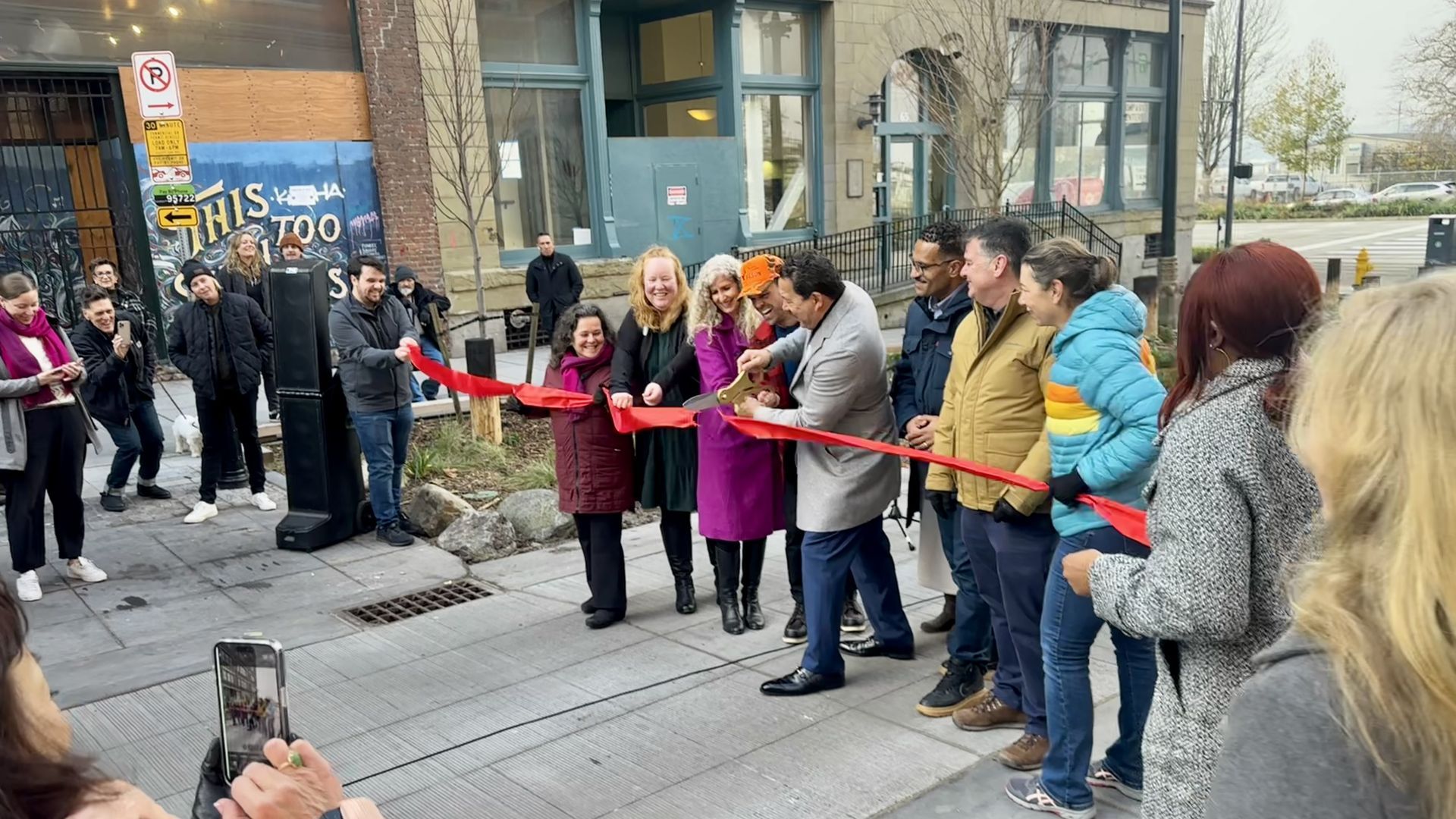 A group of Seattle officials stands behind a red ribbon while Mayor Bruce Harrell cuts the ribbon with oversized scissors.
