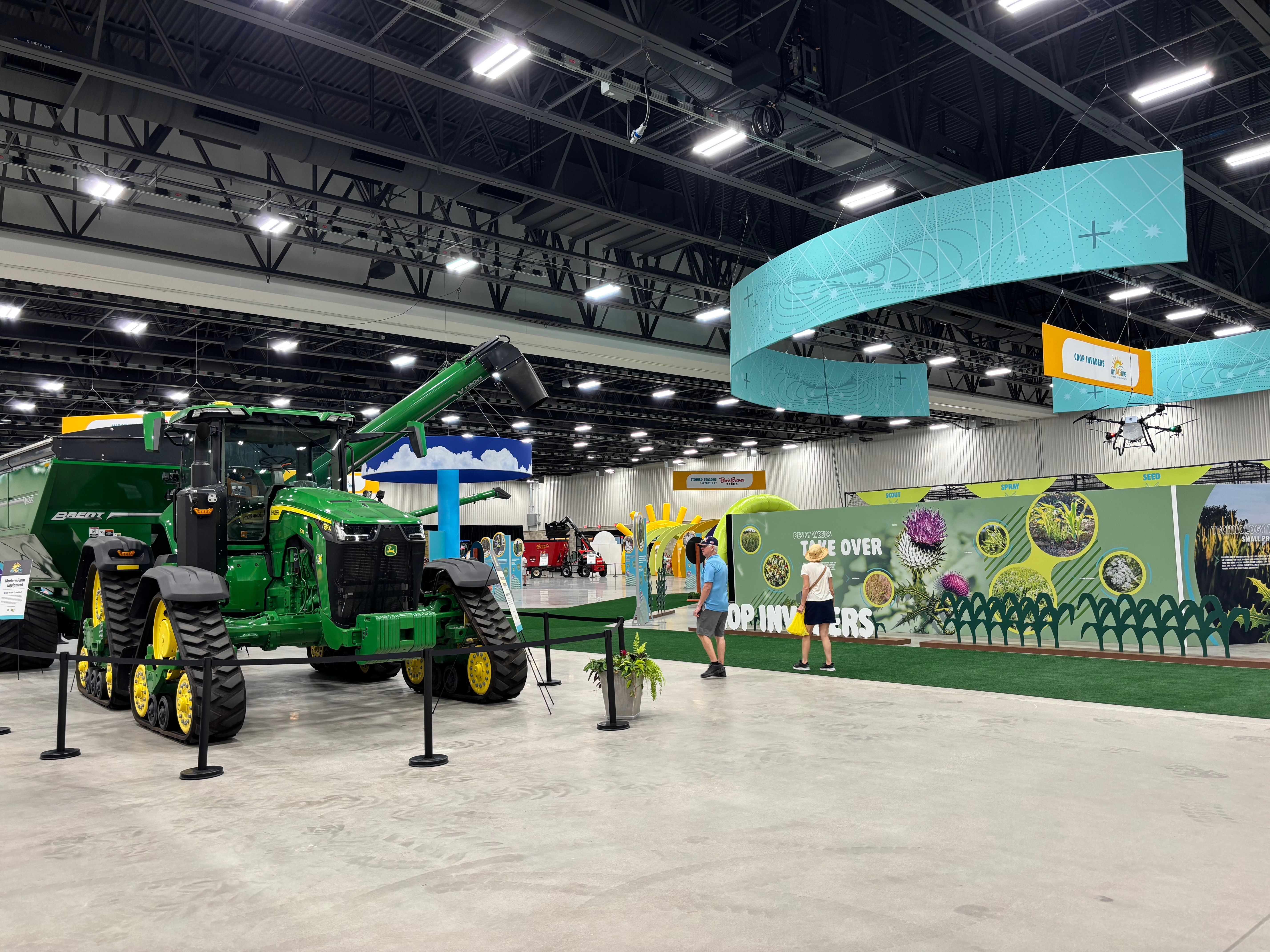 Indoor exhibition hall with a large green and yellow tractor on the left, curved teal banners overhead, and people walking past colorful farm booths and plant displays on the right.