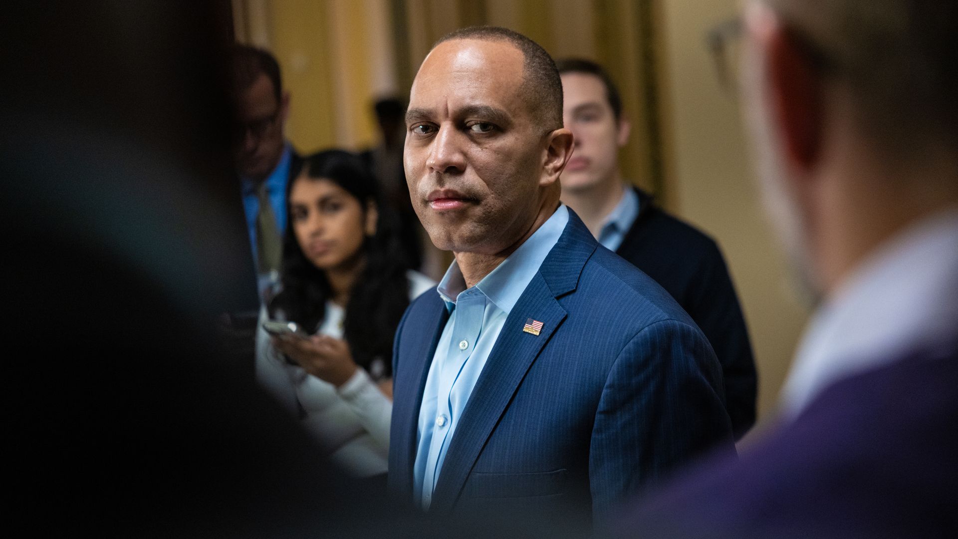 Hakeem Jeffries, wearing a blue suit, is surrounded by reporters while standing in a beige hallway.