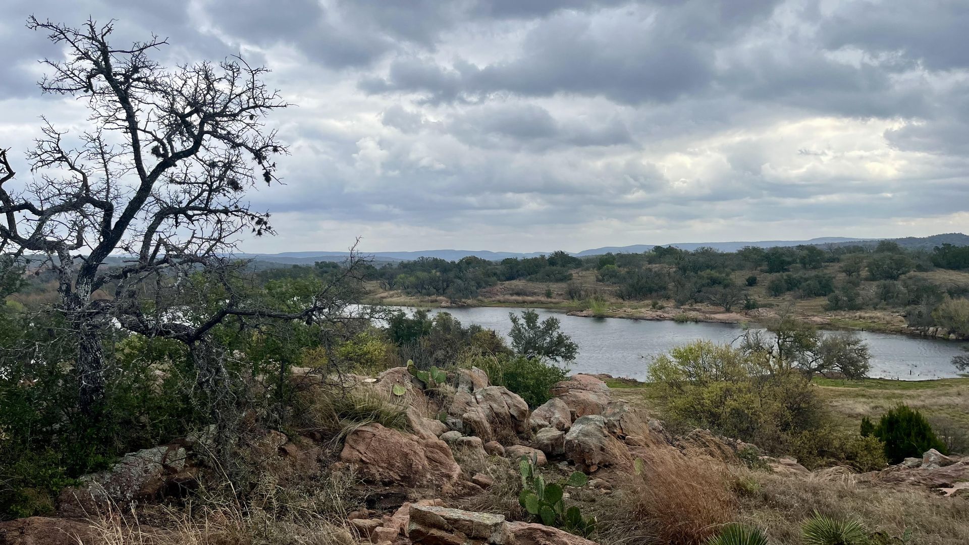 A rocky foreground with a gnarled, leafless tree on the left and dry shrubs and cacti. Beyond it sits a calm pond, with rolling hills in the distance under a cloudy sky. This is the new northern property at Enchanted Rock State Natural Area.