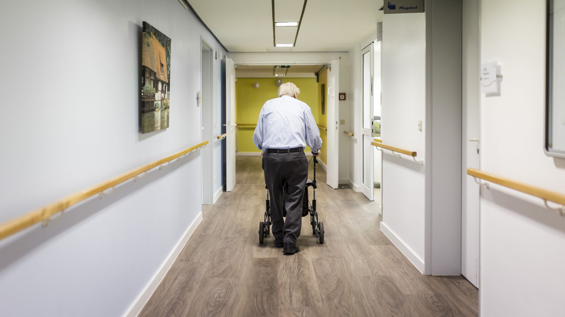 An older man walks down an empty nursing home hallway with his walker.