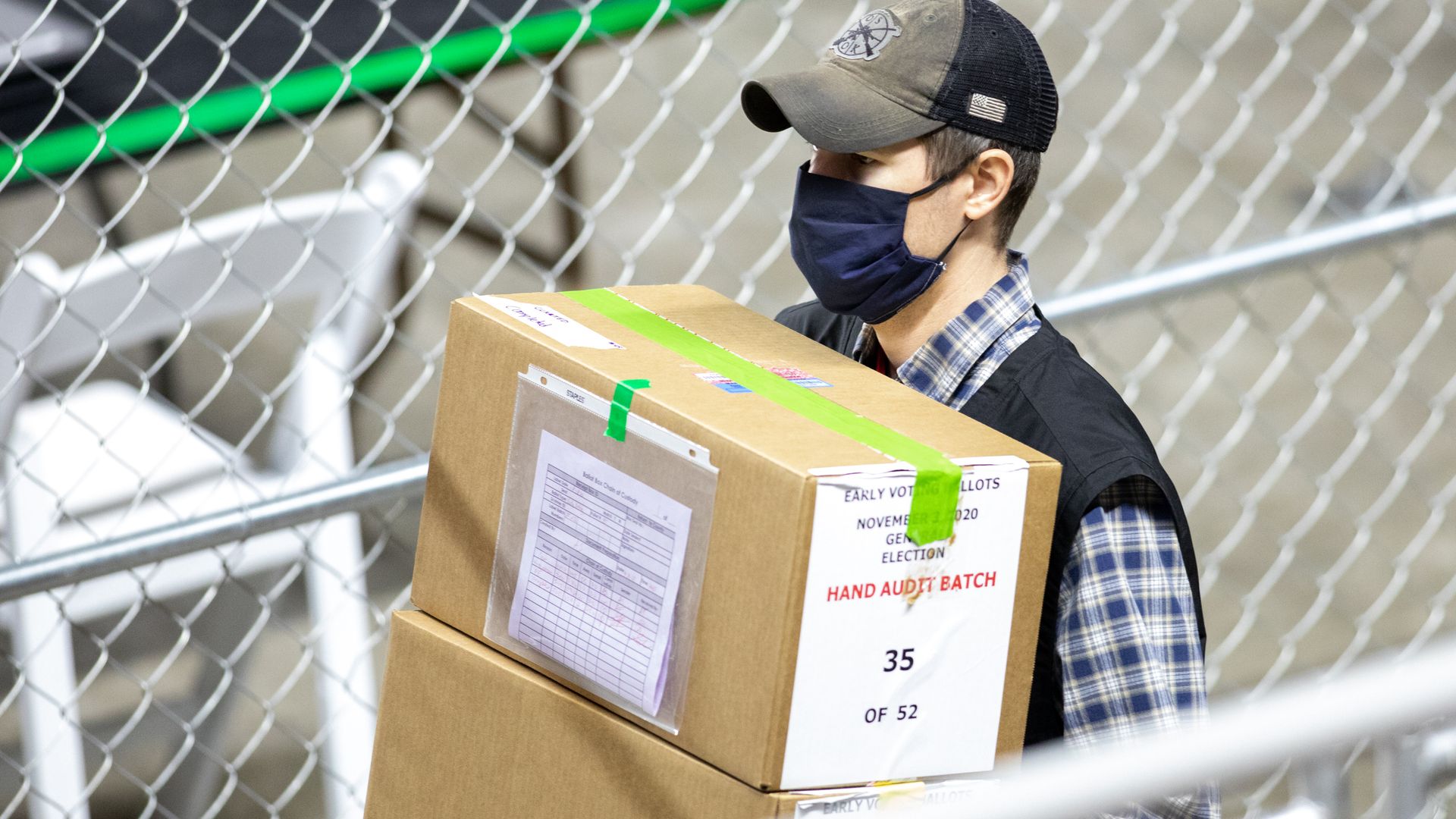 Photo of a masked worker holding two boxes of hand audit batches of votes