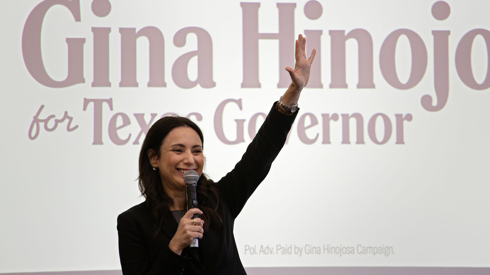 Smiling woman on stage raises her right hand while holding a microphone, in front of a large projection that reads "Gina Hinojosa" and "for Texas Governor" in text.