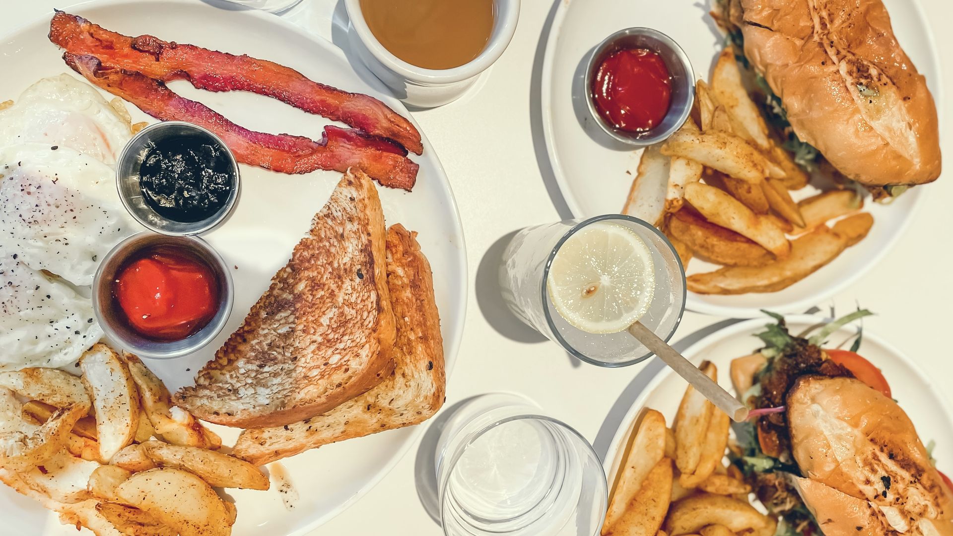 Brunch food including bacon, fries and sandwiches on a bright white table.