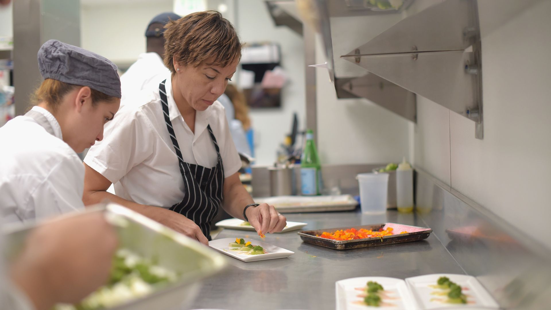 Chef Nina Compton plates a dish inside a busy kitchen. Two other chefs are seen beside her working.