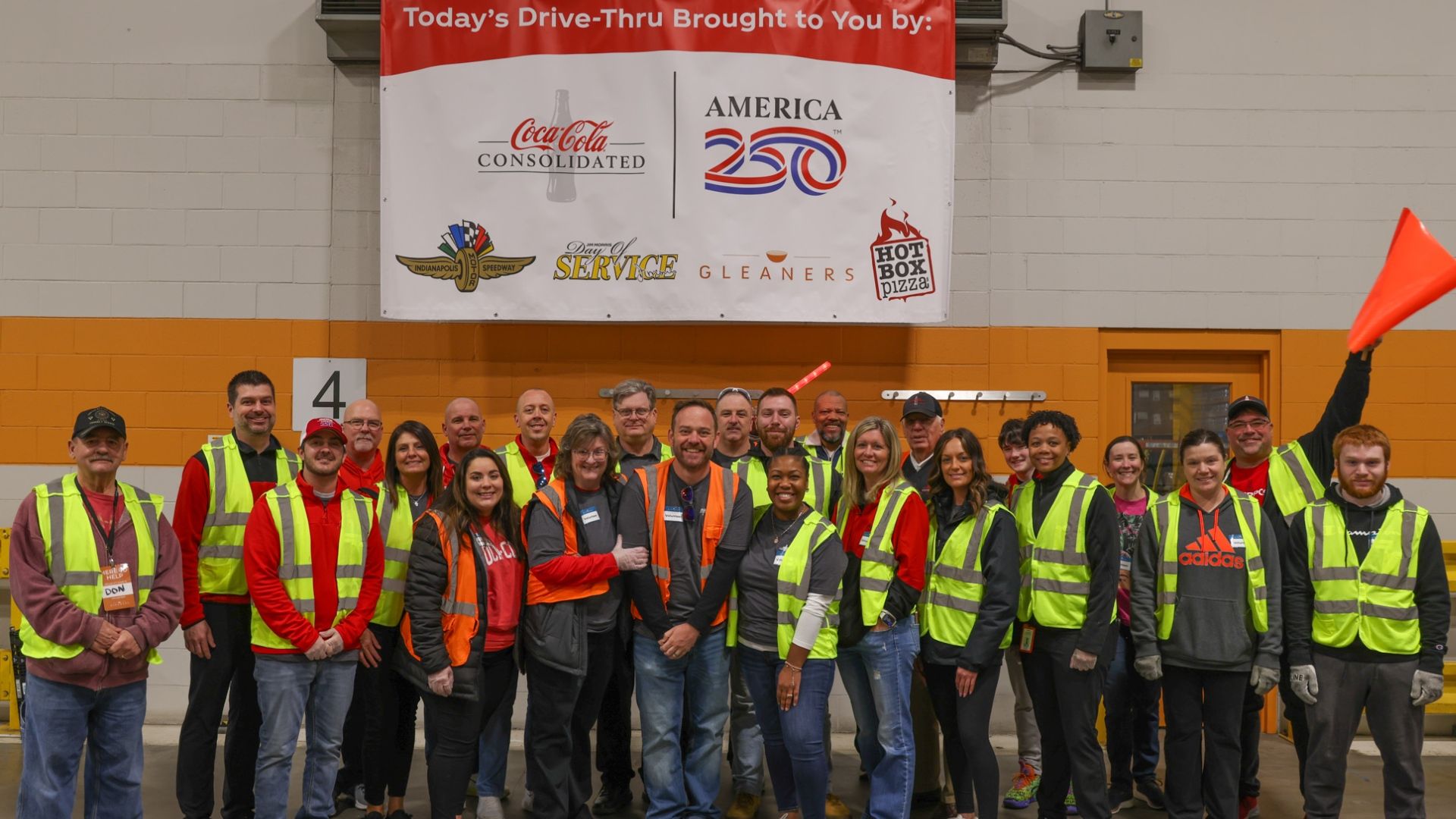 A large group of volunteers in neon safety vests pose for a photo indoors beneath a banner featuring Coca-Cola Consolidated, America 250, Indianapolis Speedway, Day of Service and other logos.
