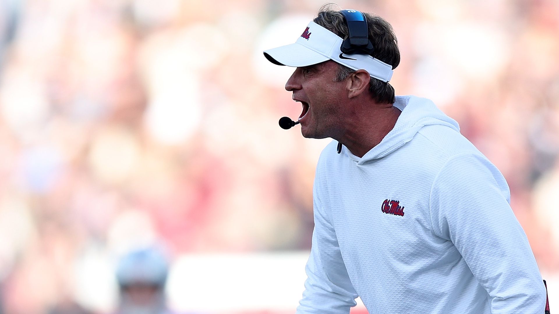 A man is photographed mid-yell. He wears a white Ole Miss hoodie and visor.