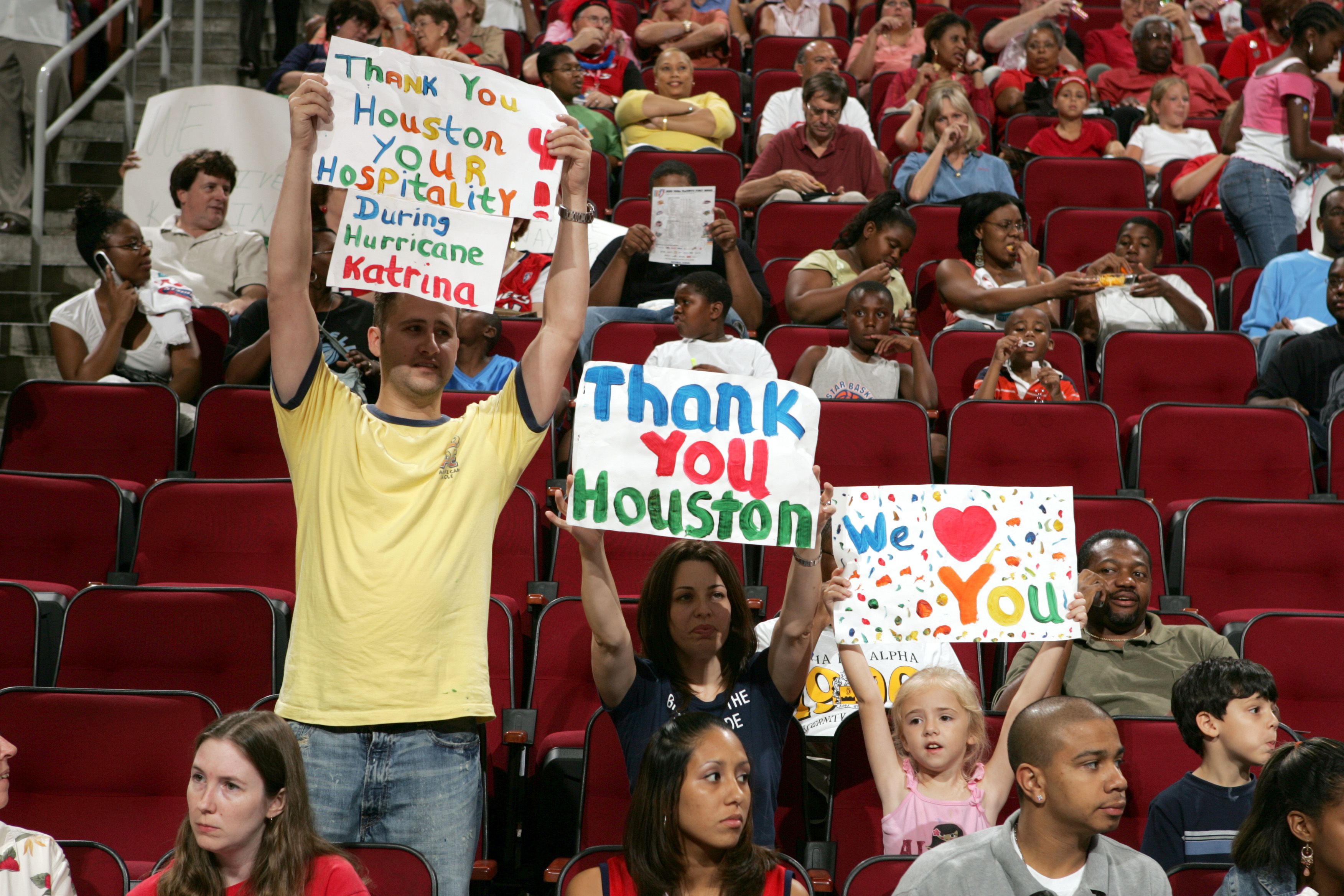 A group of people seated in red stadium chairs hold colorful handwritten signs thanking the city of Houston for hosting evacuees from New Orleans