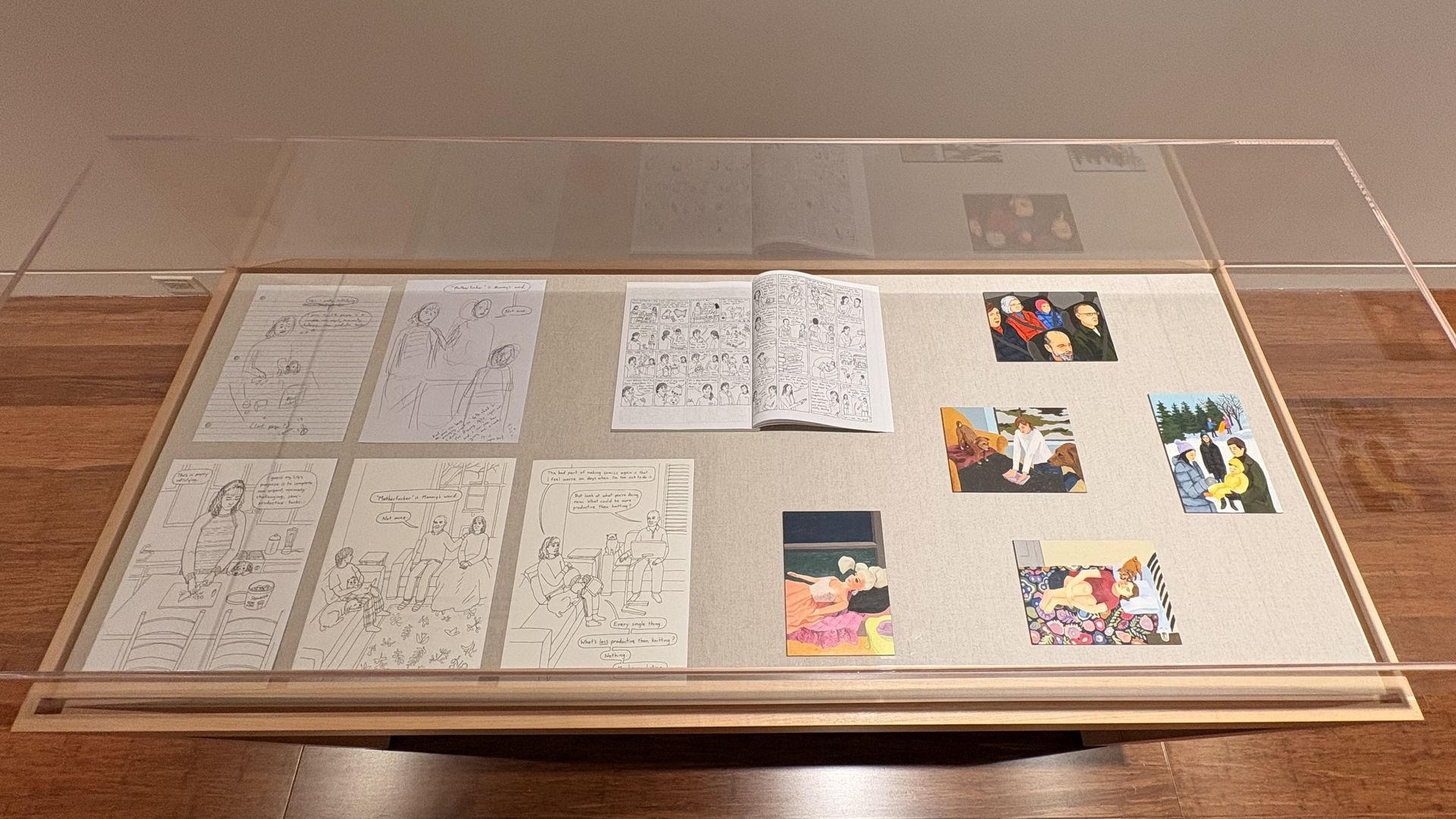 A glass display case on a wooden floor, showing open comic pages and small color panels laid out on a beige fabric background, with a plain beige wall behind.