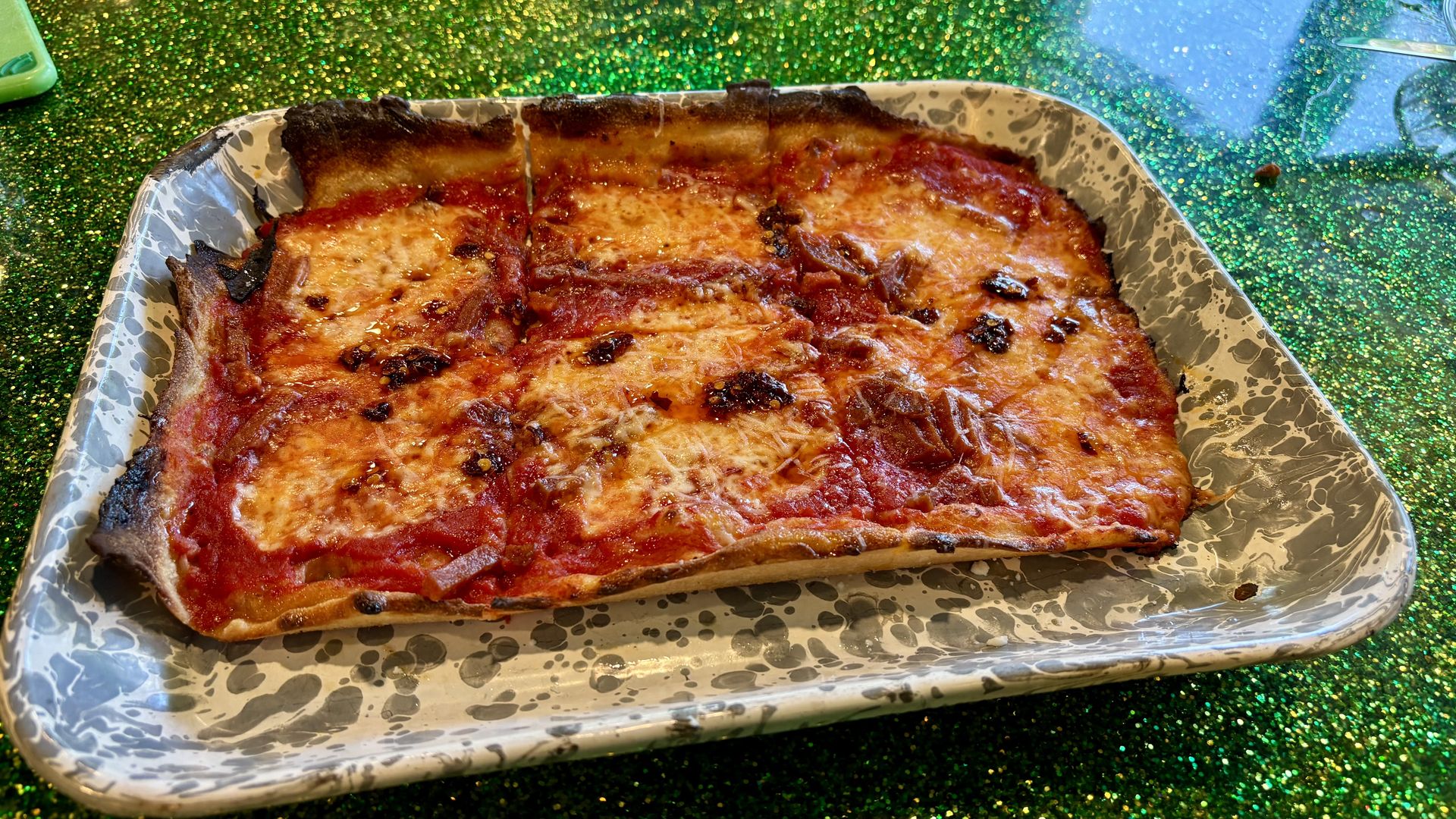 Rectangular cheese pizza with slightly burnt edges on a gray and white speckled metal tray, resting on a sparkly green surface.