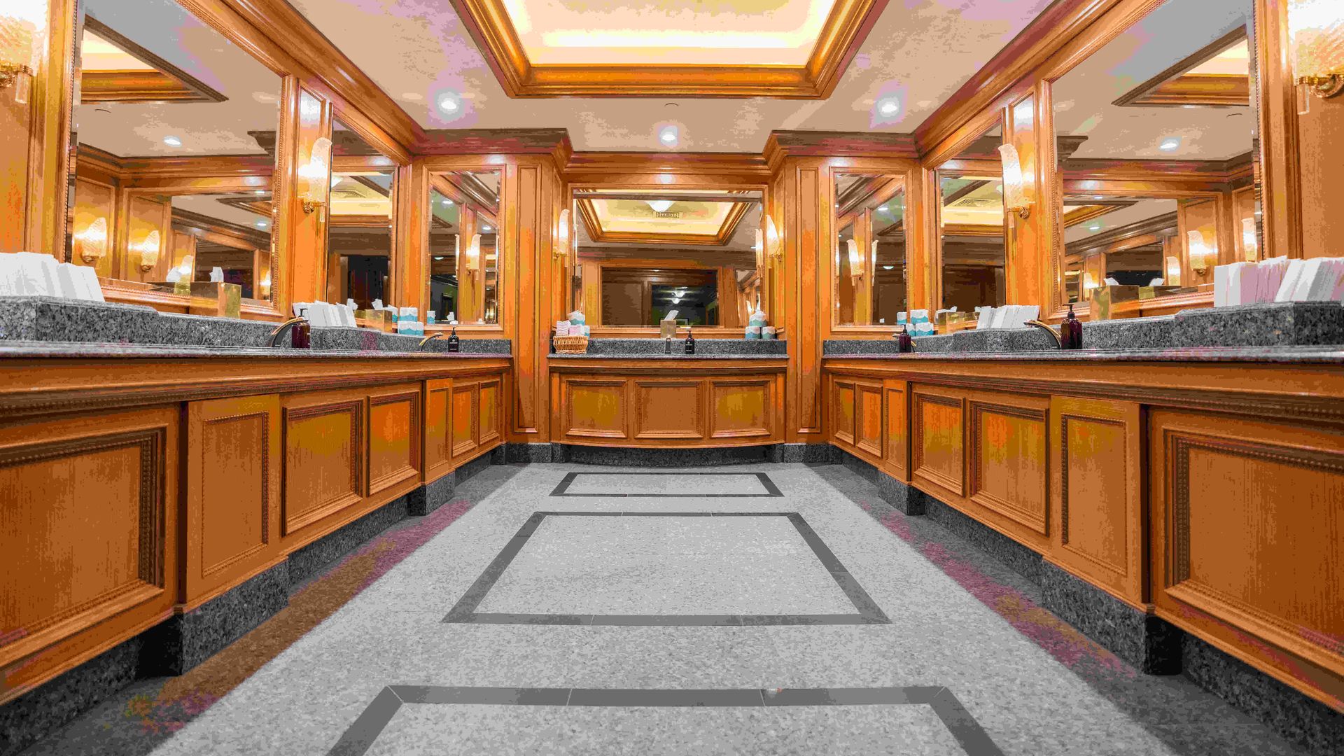 The sink area in a public bathroom, with mirrors, wood molding and sconces.