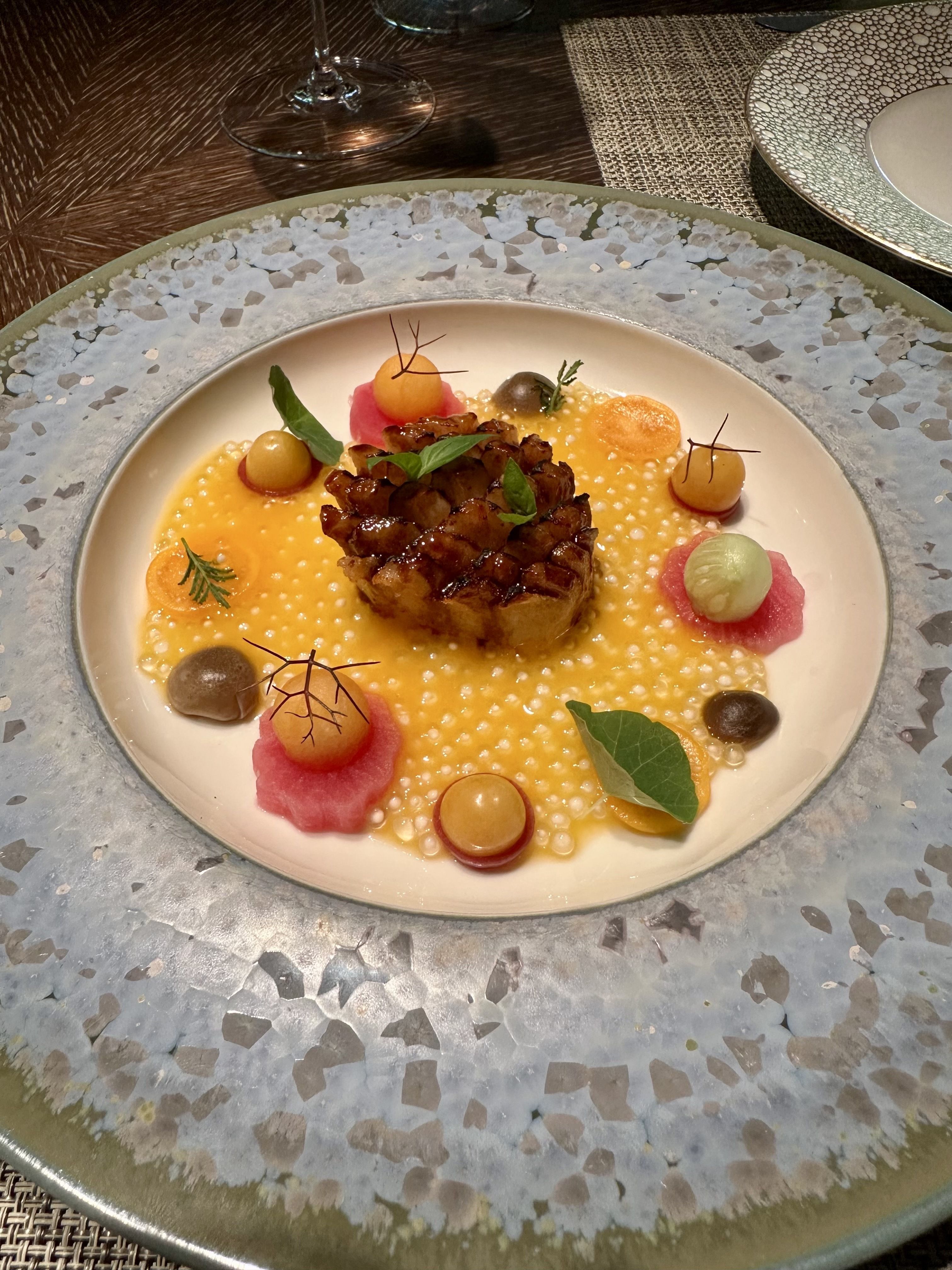 Elegant plated dish on a textured charger: caramelized meat centerpiece with yellow sauce, small round quenelles, greens, and colorful garnish; wine glass and patterned plate in background.