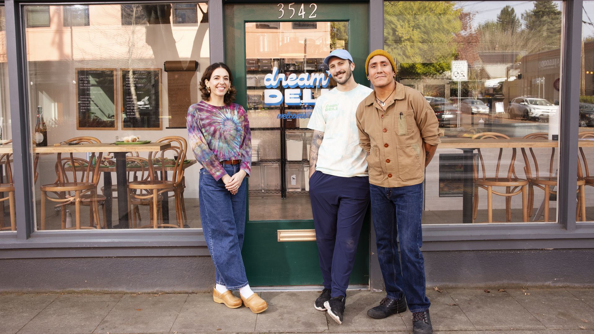 Three smiling people standing outside a green-door deli with the sign "dream DELI". The woman wears a tie-dye top and jeans, the men wear a white shirt and beige jacket respectively.